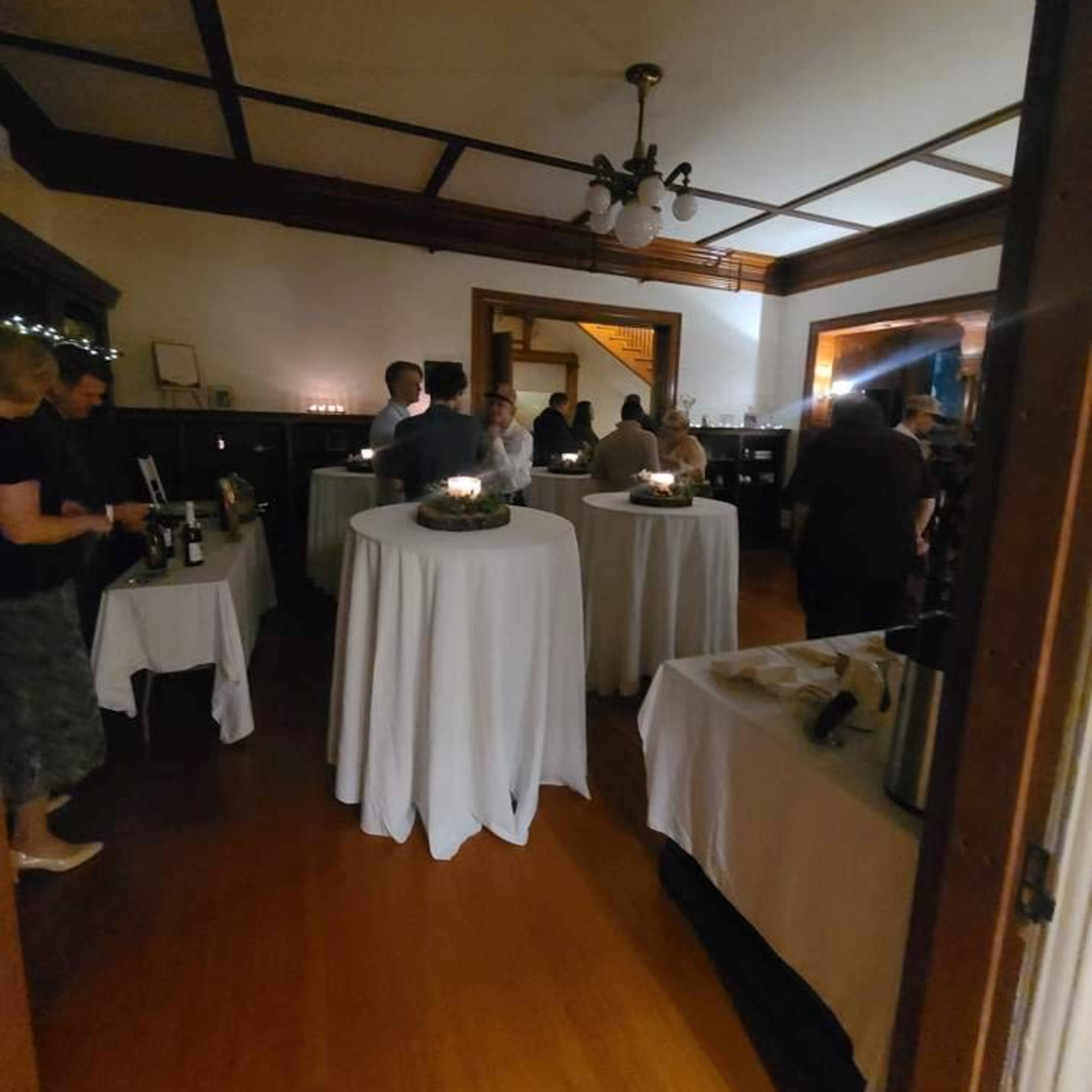 A group of people socializes in a warmly lit room filled with round tables covered in white tablecloths.