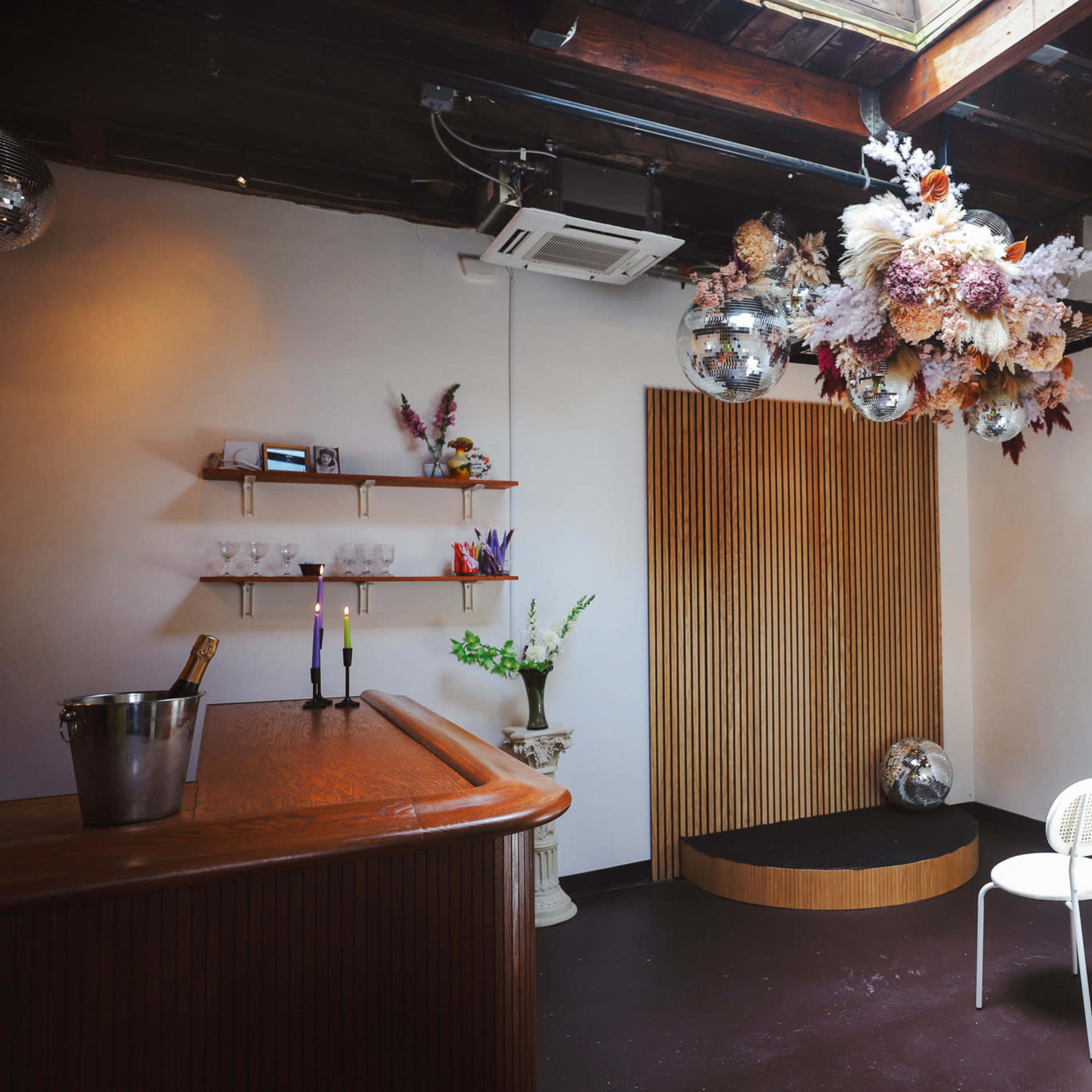 A stylish interior space featuring a wooden bar, a flower and disco ball installation overhead, and a small stage area with white chairs arranged in front of it.