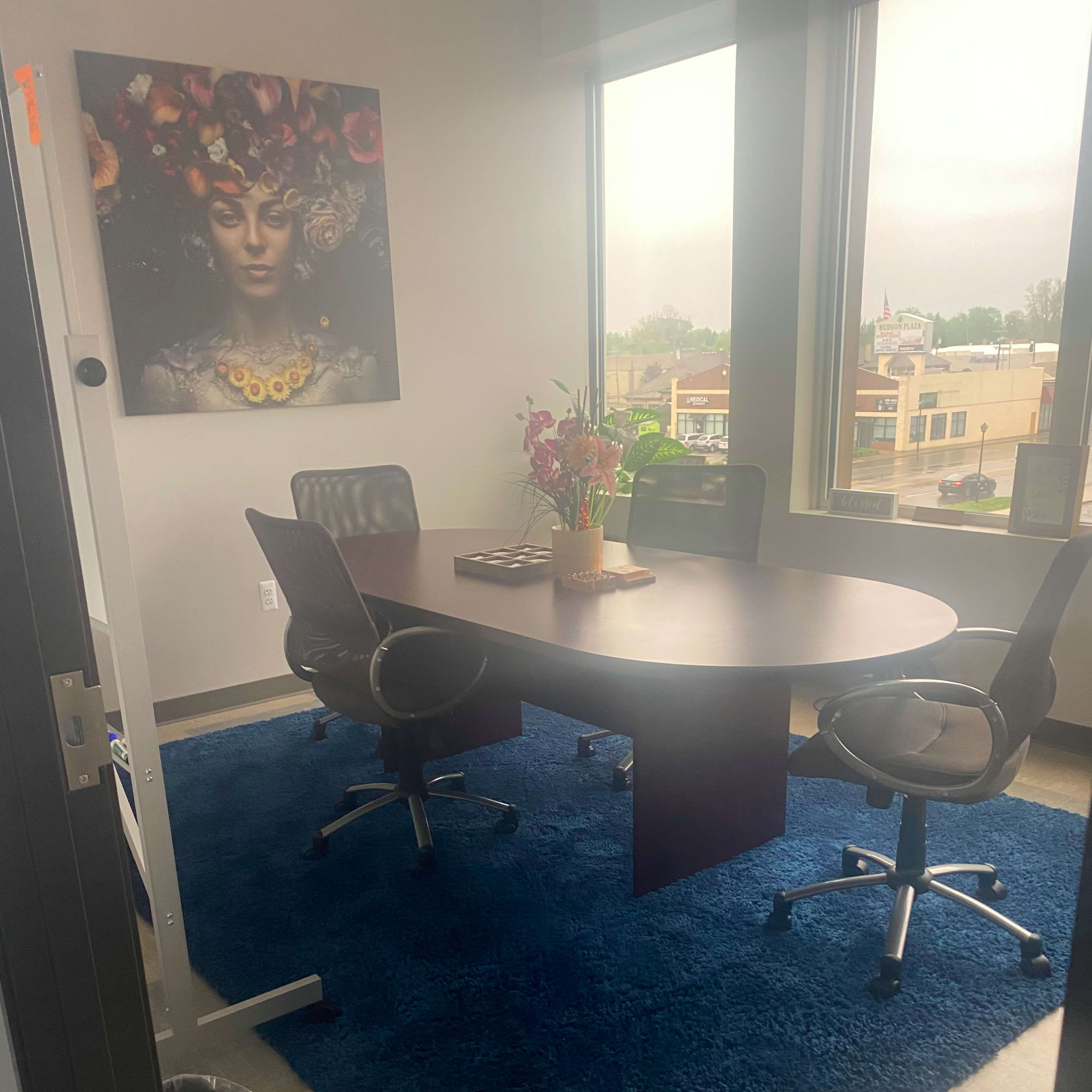 A conference room with a round table surrounded by four chairs, featuring a large window, a colorful wall art piece, and a rug on the floor.