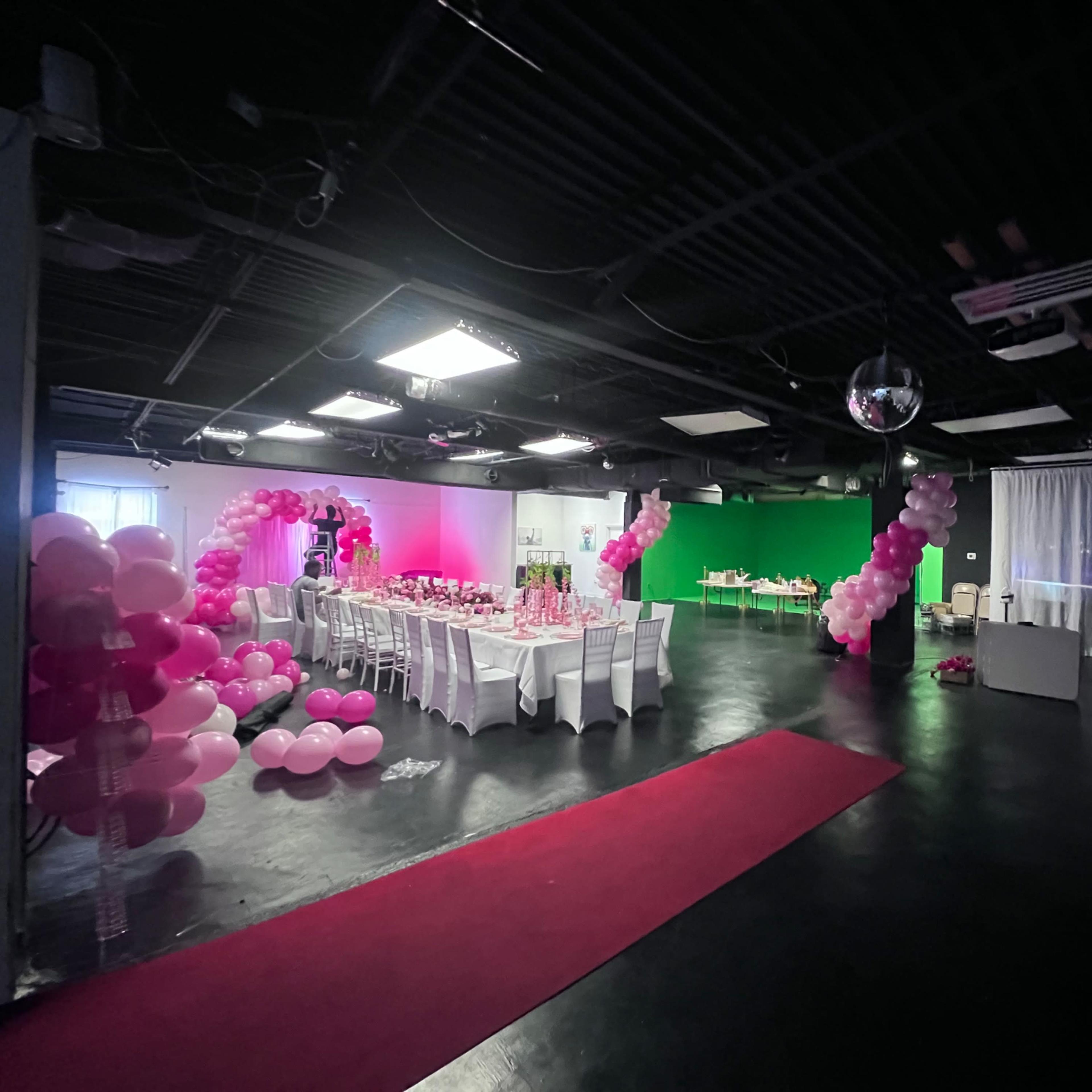 An event space decorated with pink balloons, a red carpet, and a green backdrop, featuring tables set for a gathering on one side and a projection of a figure on the wall.