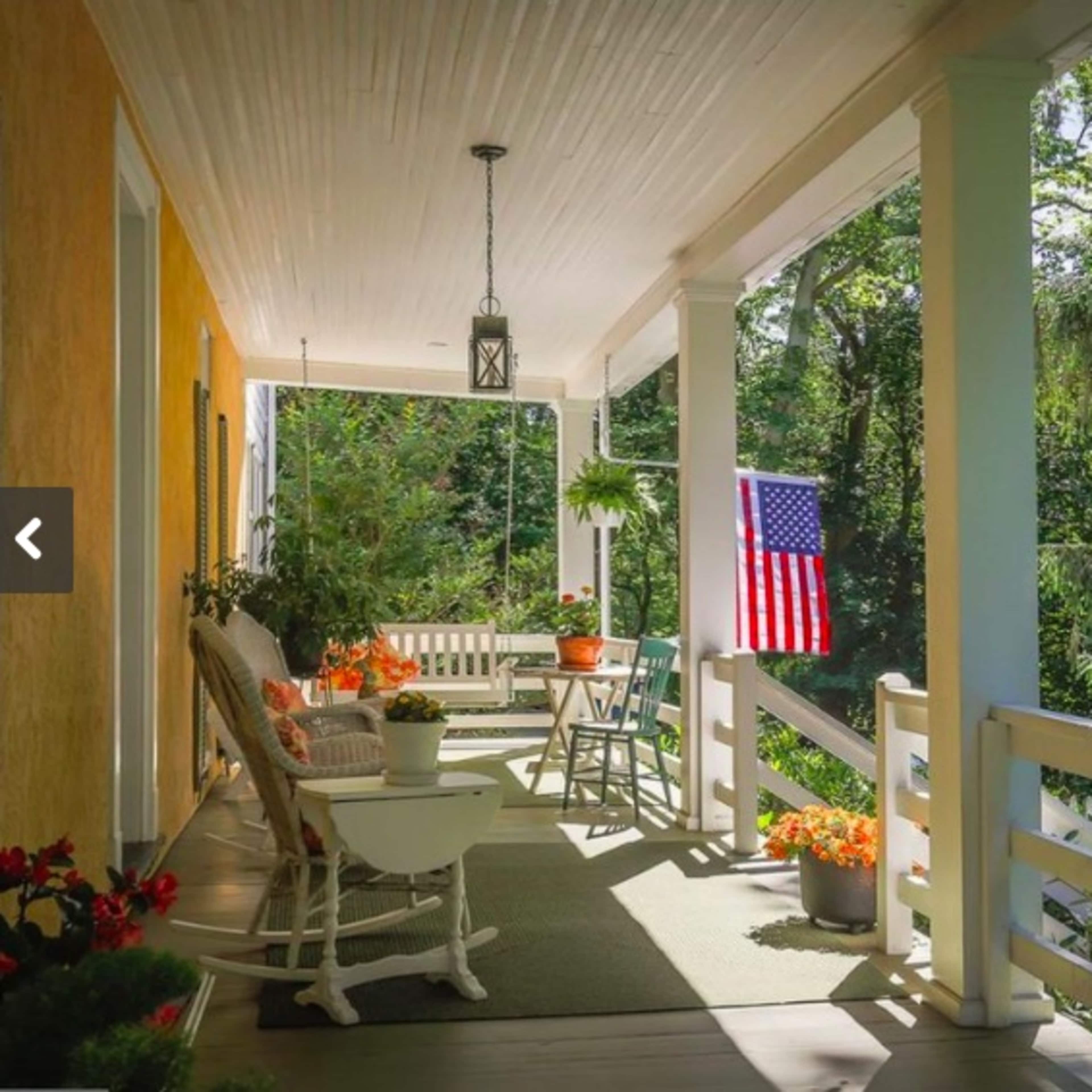 The porch features a seating area with rocking chairs, a small table, and an American flag hanging amidst lush greenery.