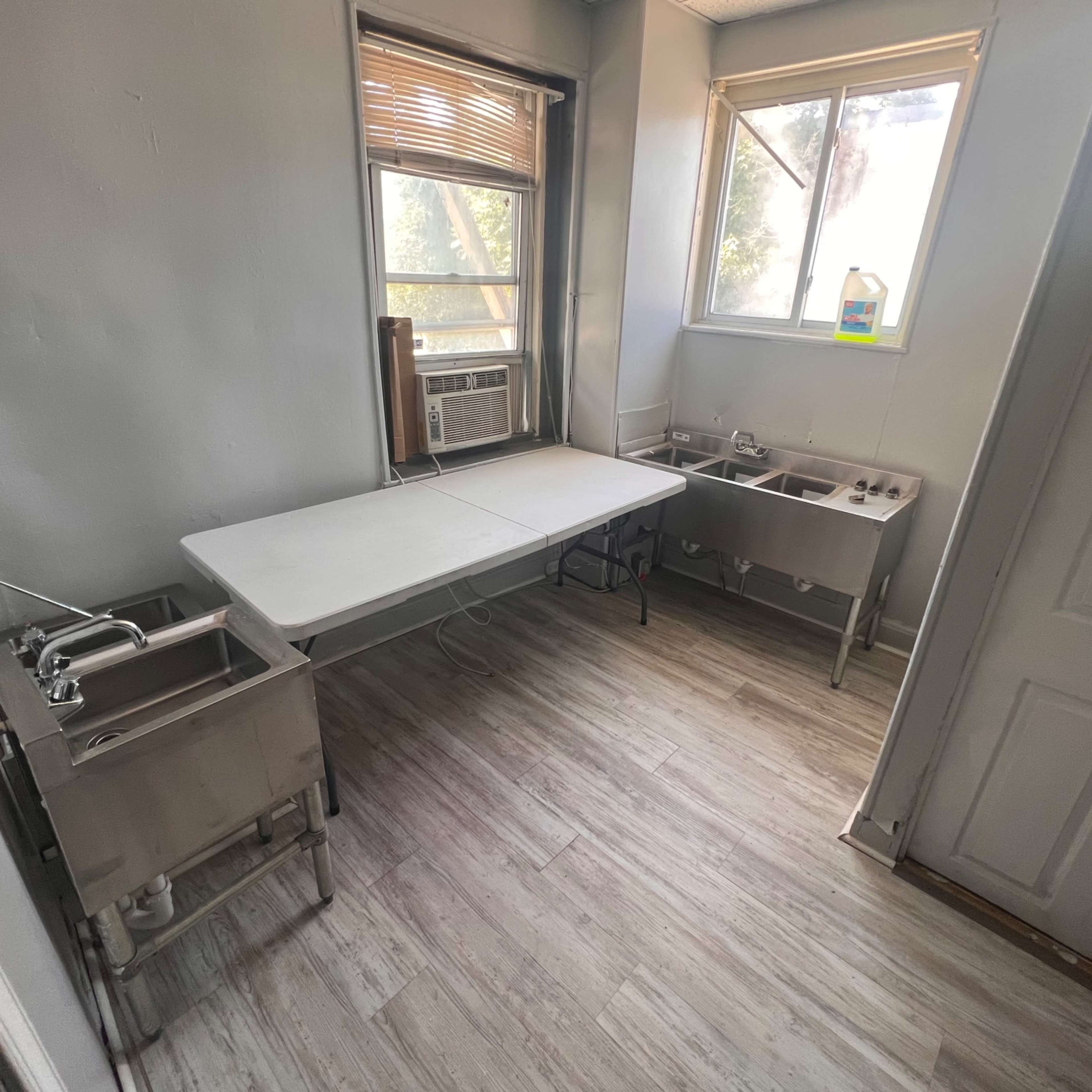 A small kitchen space featuring a table in the center, two metal sink units on the right, and a window on the back wall.