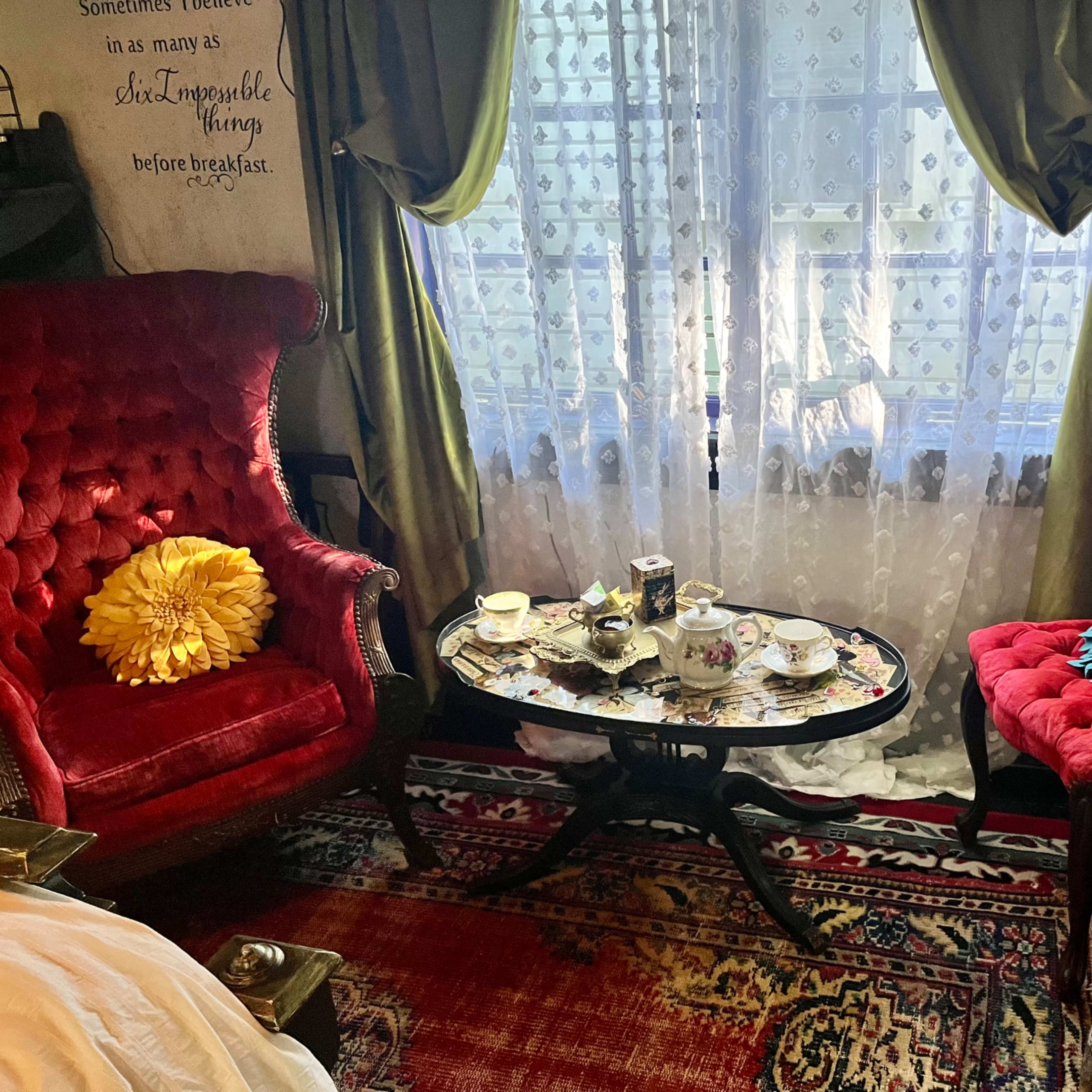 The scene depicts a quaint seating area with two red velvet chairs, a decorative coffee table covered in tea set items, and a window adorned with lace curtains.