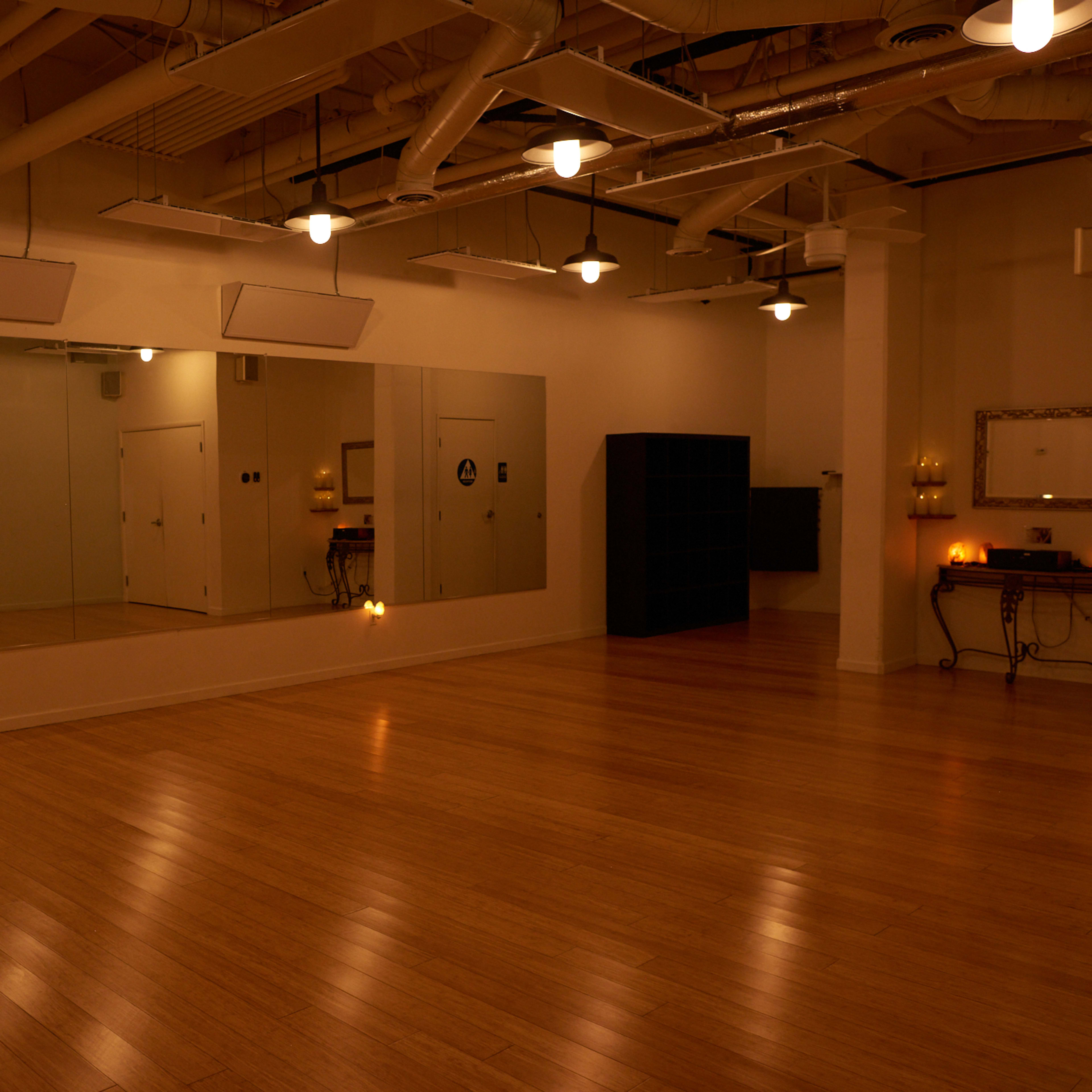 A spacious studio with hardwood flooring and mirrors along one wall, illuminated by overhead lights.