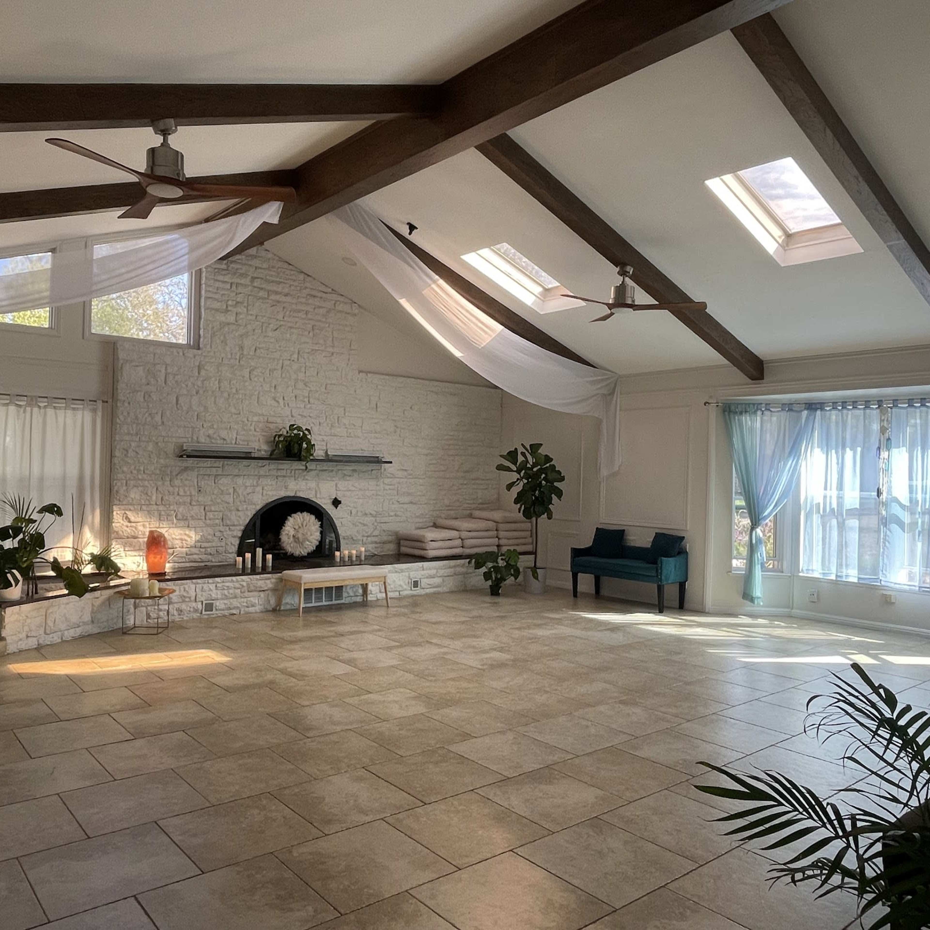 A spacious, well-lit room with high ceilings, large windows, and a stone fireplace, furnished with potted plants and a blue sofa.