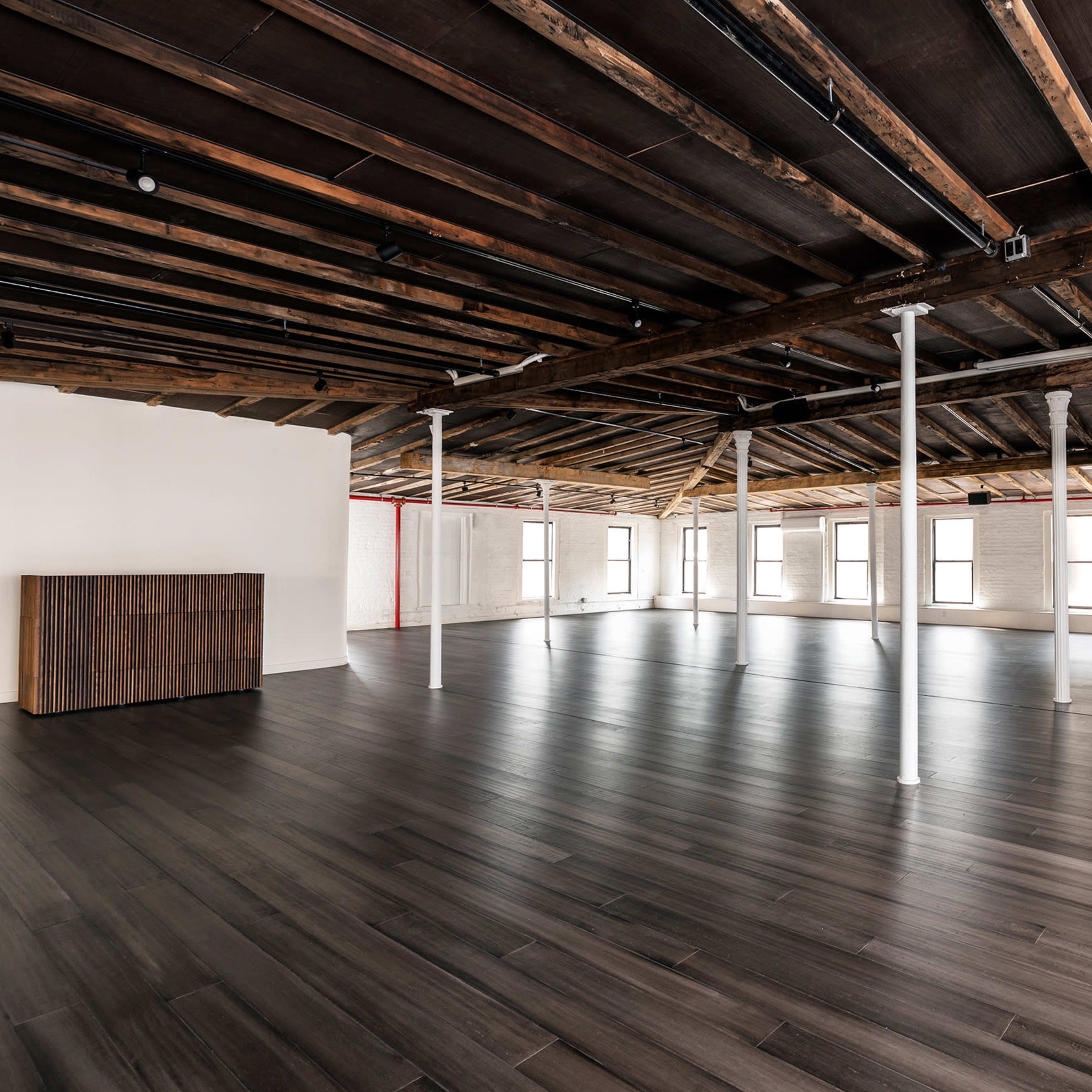 A spacious, empty room with wooden beams and a polished floor, featuring multiple windows along the walls.