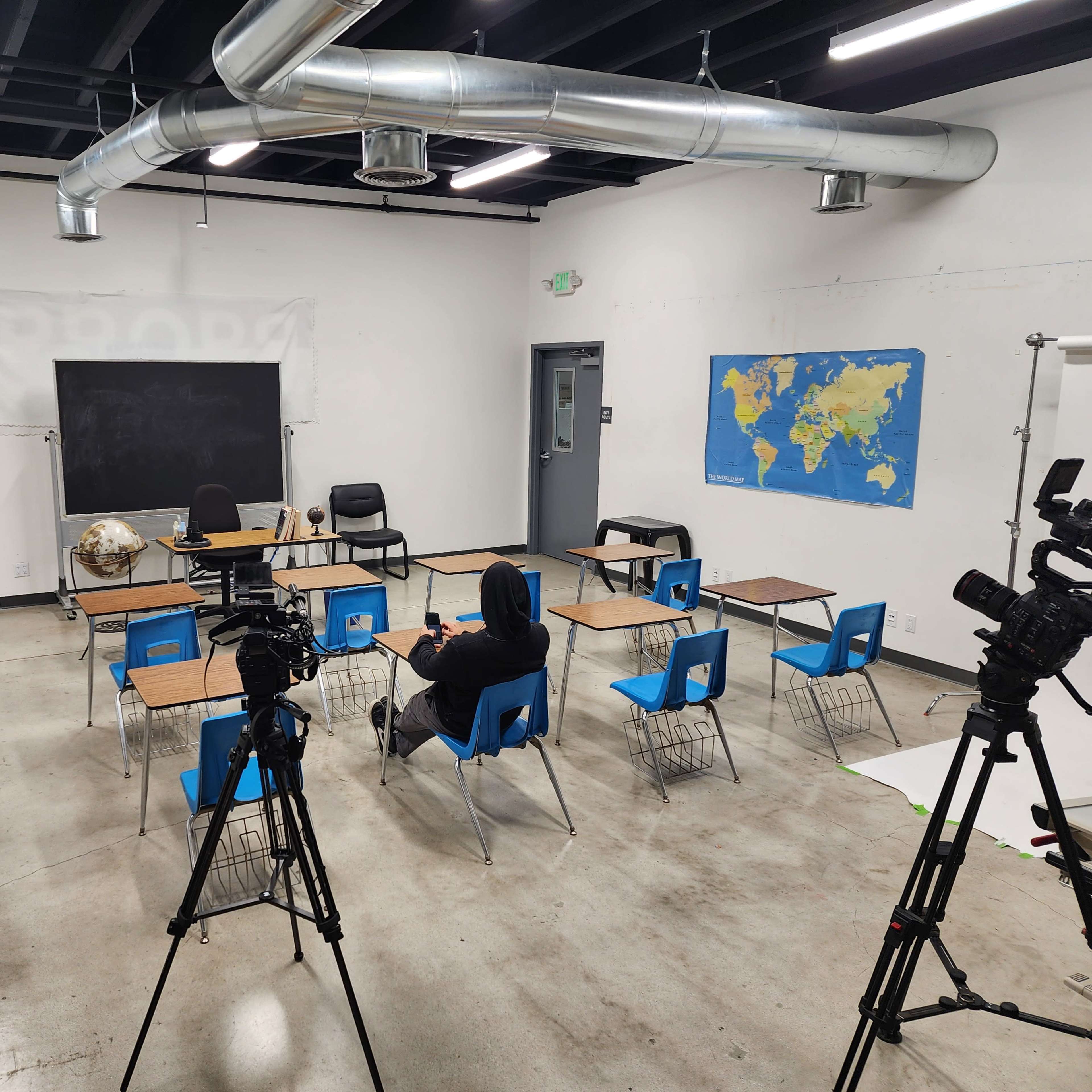 An empty classroom setup with several desks and chairs arranged around a central space, flanked by cameras on tripods.