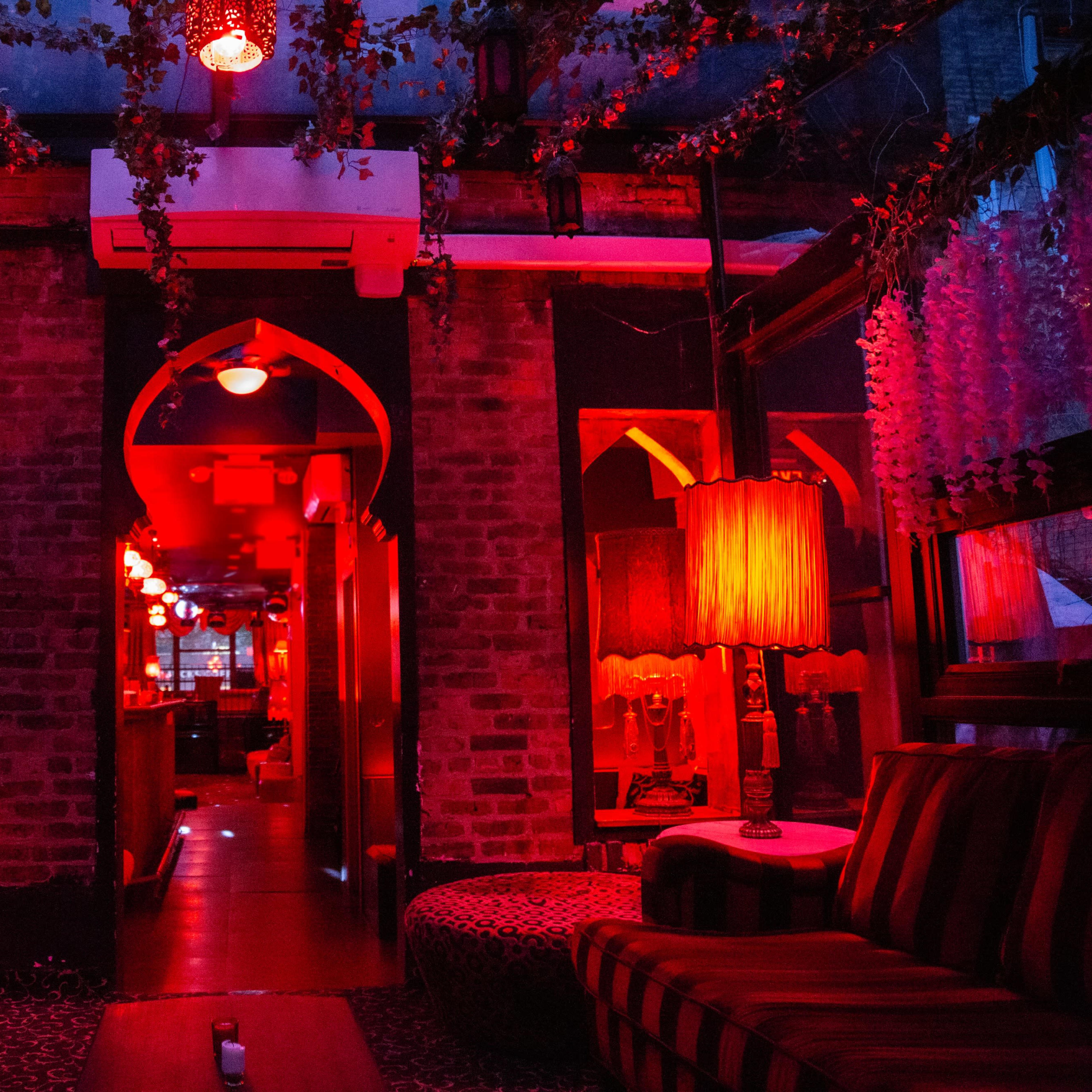 A dimly lit lounge with red lighting, plush seating, and decorative lamps lining the walls, leading to a doorway at the back.