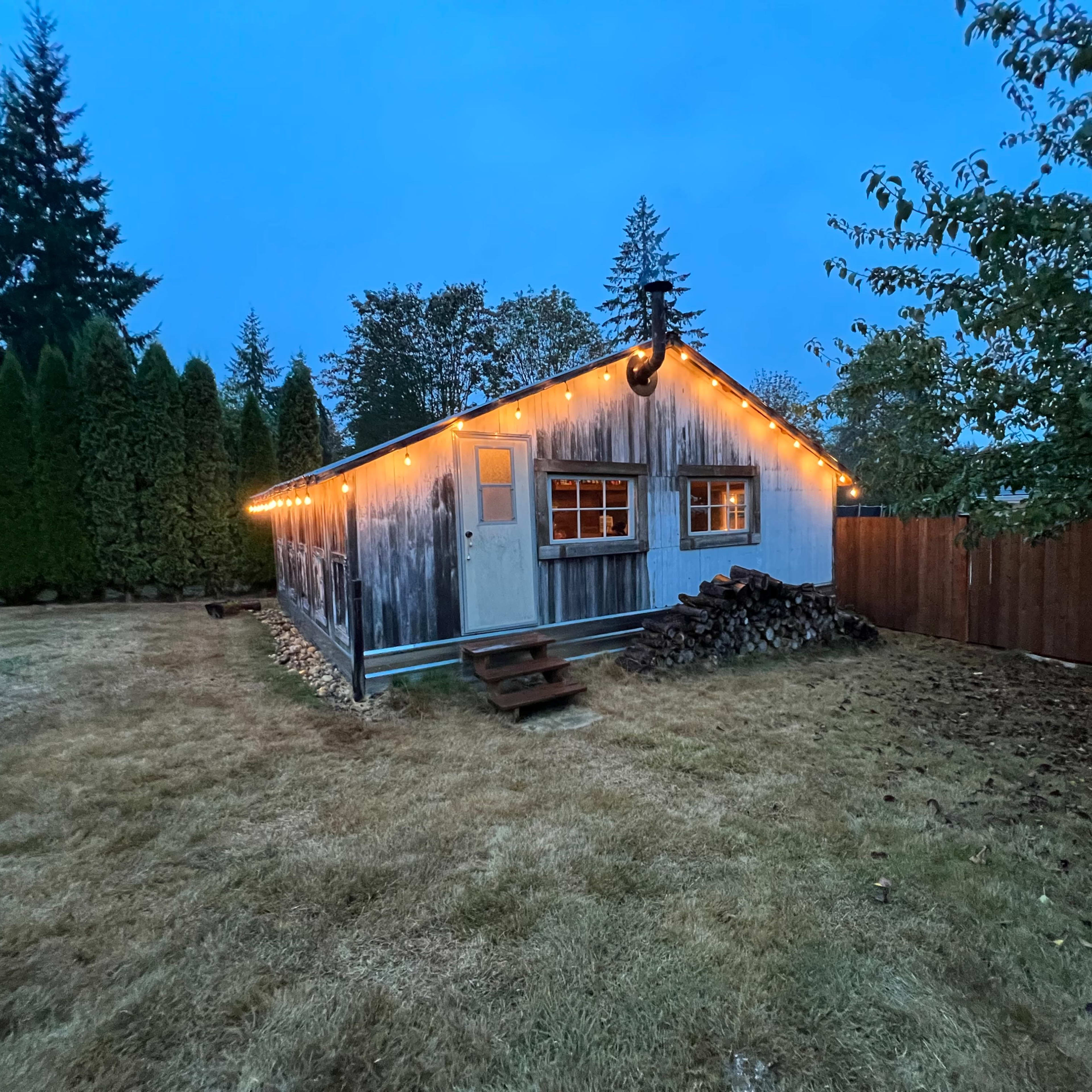 A quaint cabin with string lights is set in a grassy yard surrounded by tall trees and a fenced area.