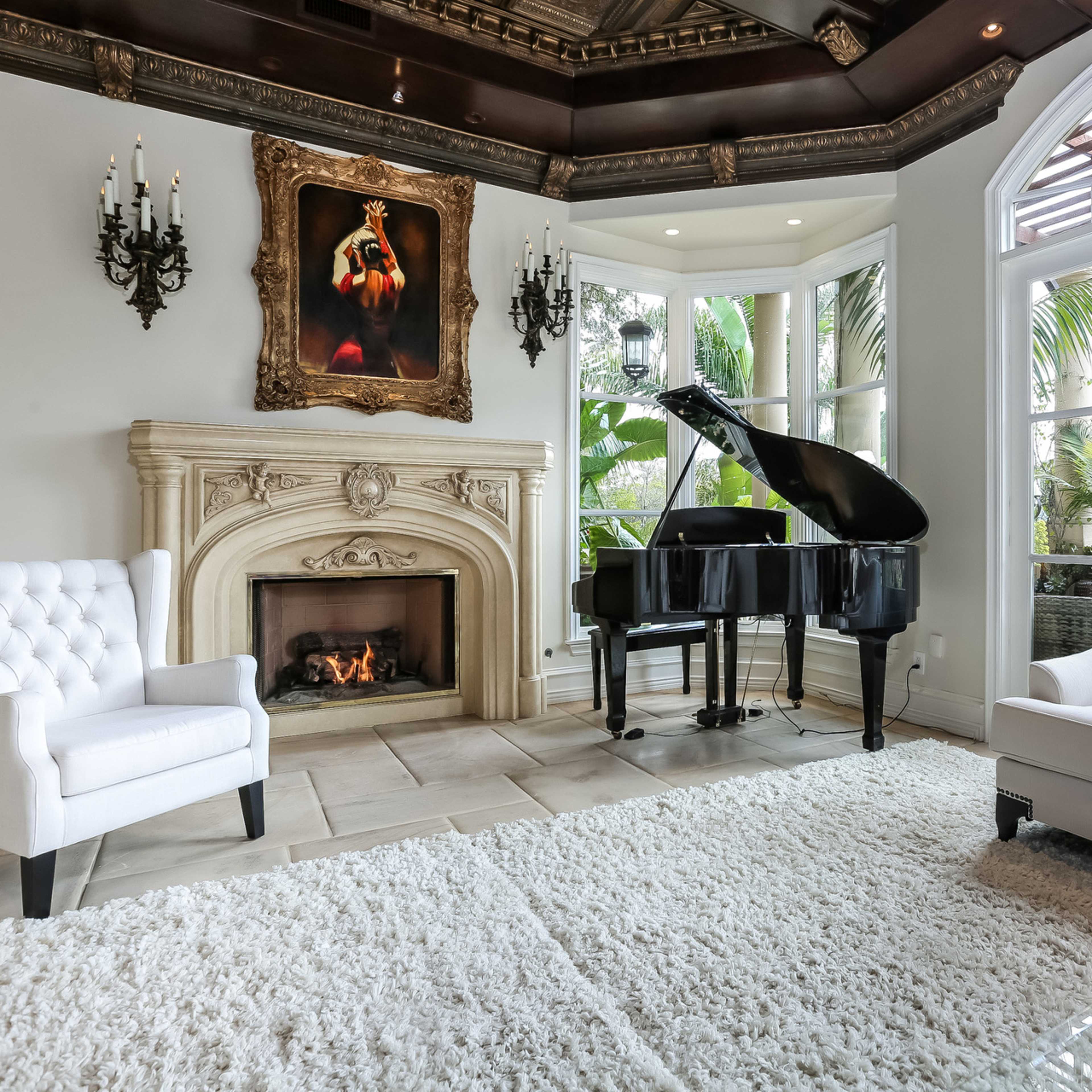 A living room features a grand piano, two white armchairs, a fireplace, and large windows overlooking greenery.