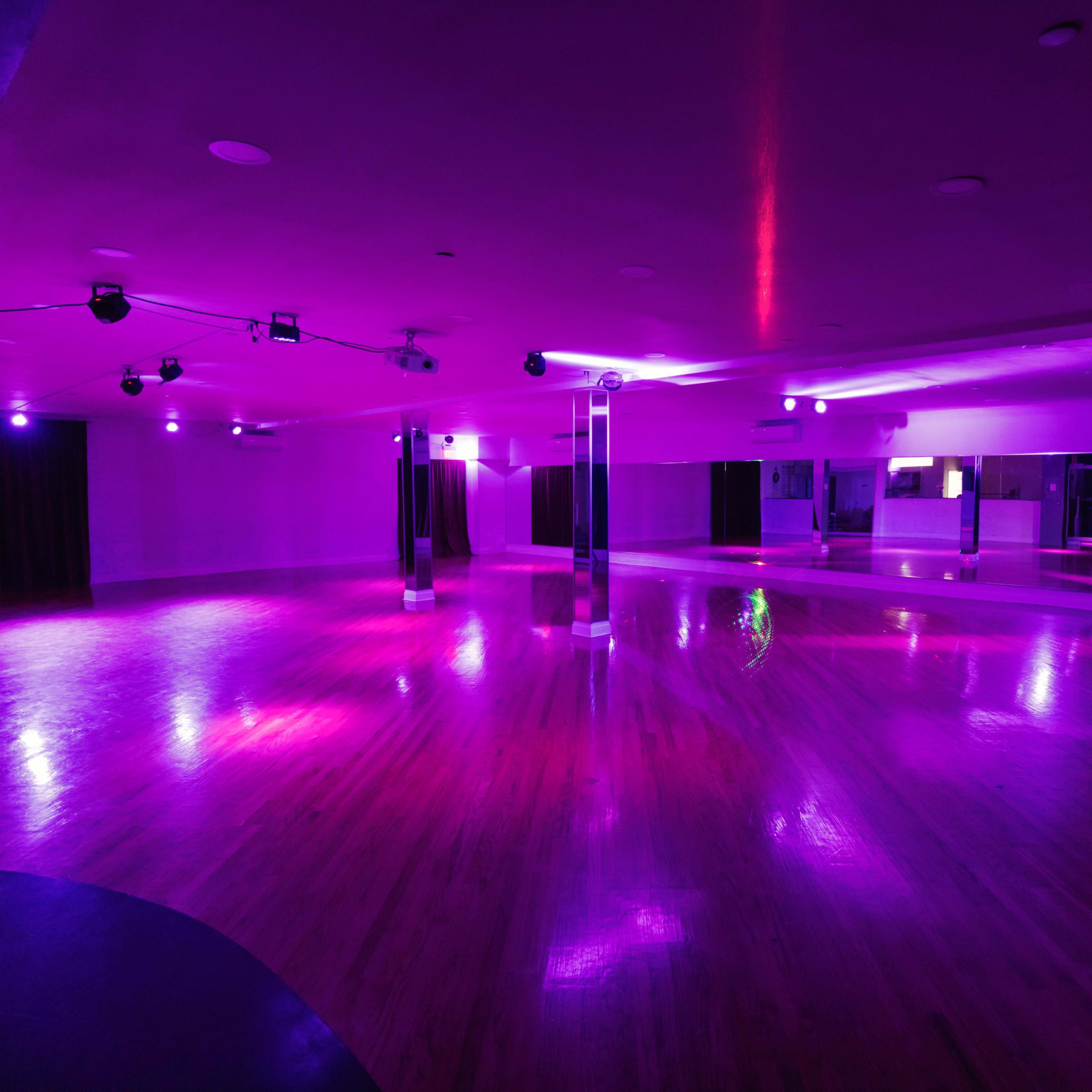 A dance studio with polished wood floors and purple lighting, featuring mirrors along one wall.