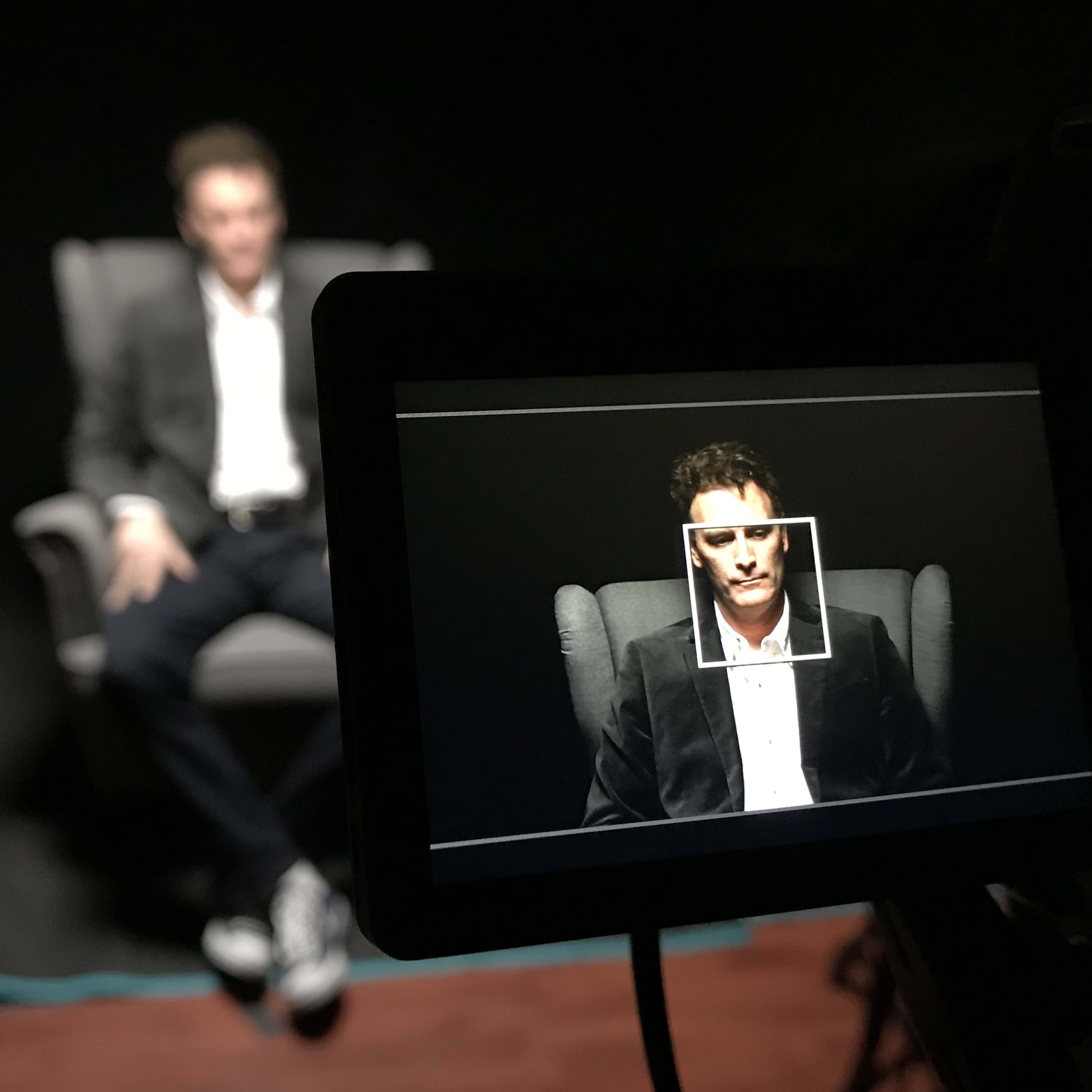 A man sits in a chair, framed within a camera's display, while the background is dark.