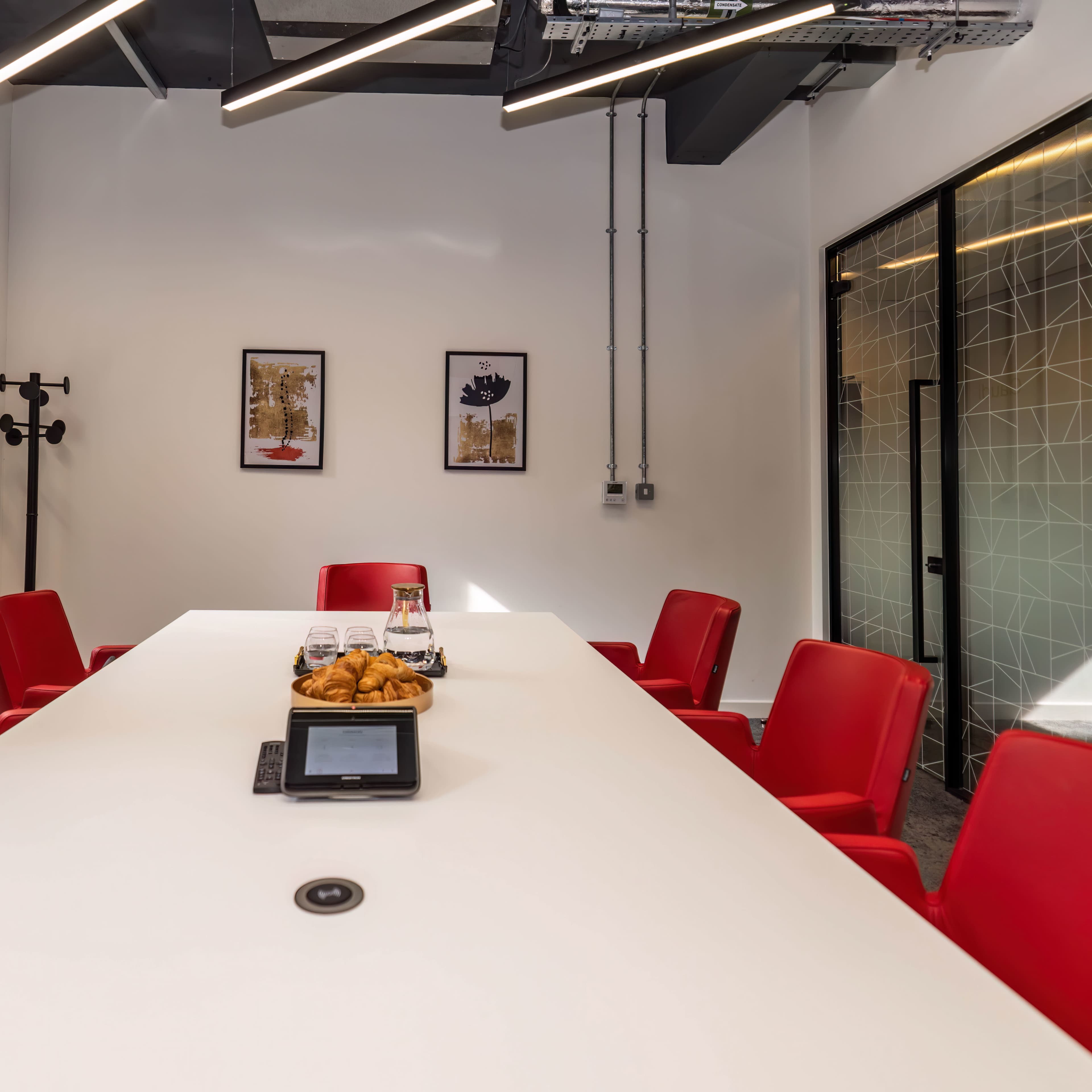 A modern conference room features a white table surrounded by red chairs, with framed artworks on the wall and a tray of pastries on the table.