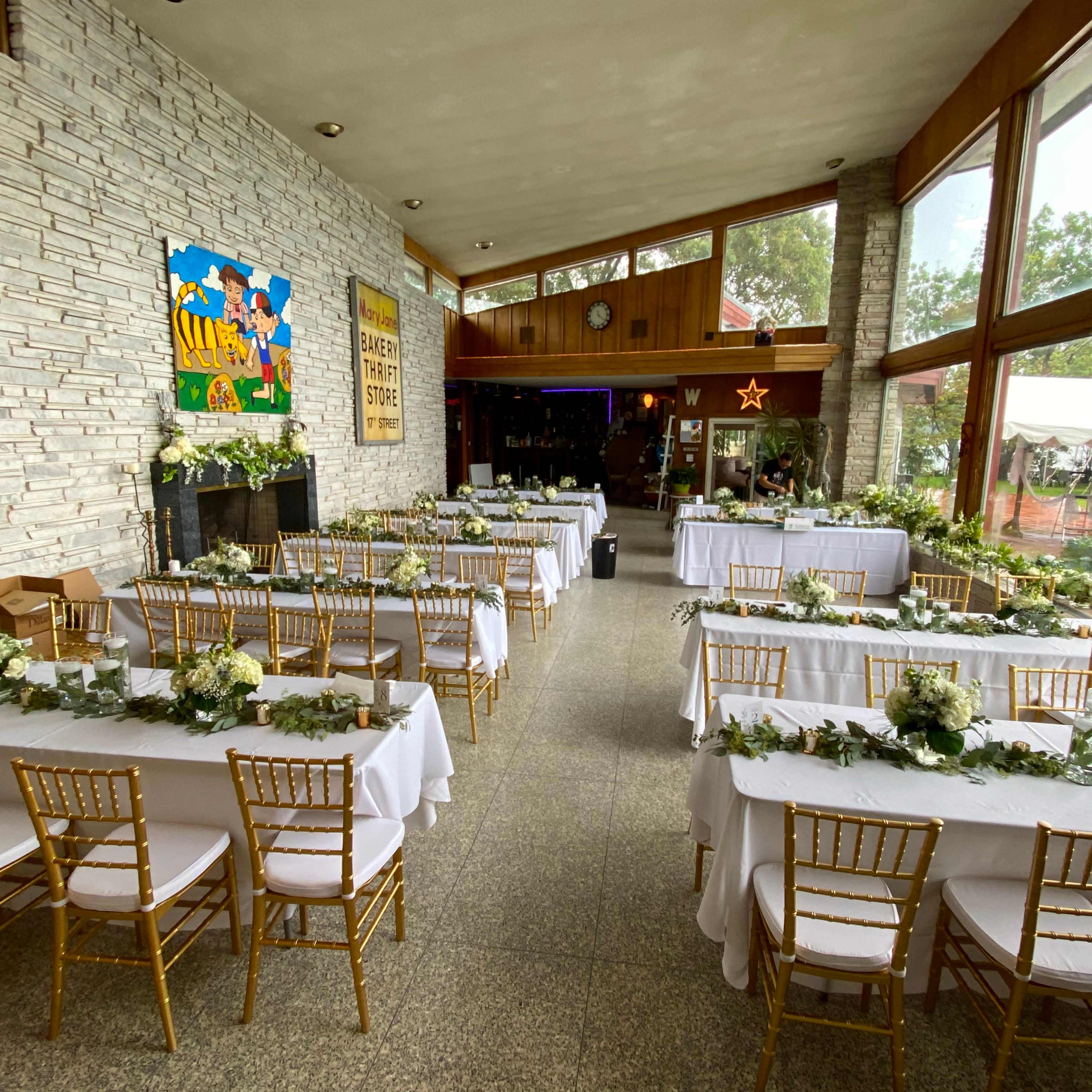 A decorated event space with tables arranged in rows, adorned with white tablecloths and floral centerpieces, set for a gathering or reception.