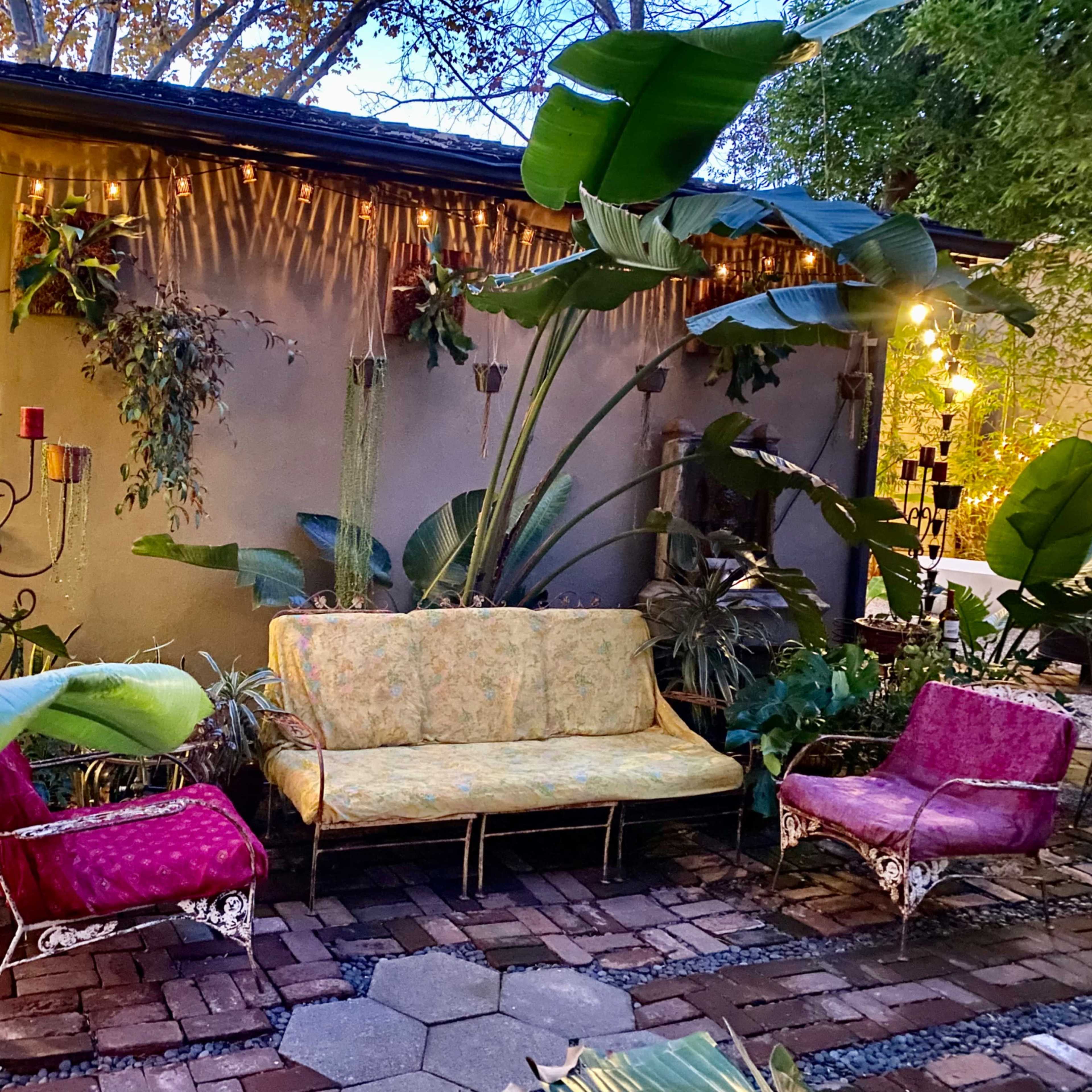 A cozy outdoor seating area features a floral-patterned couch and two colorful chairs surrounded by tropical plants and soft lighting.