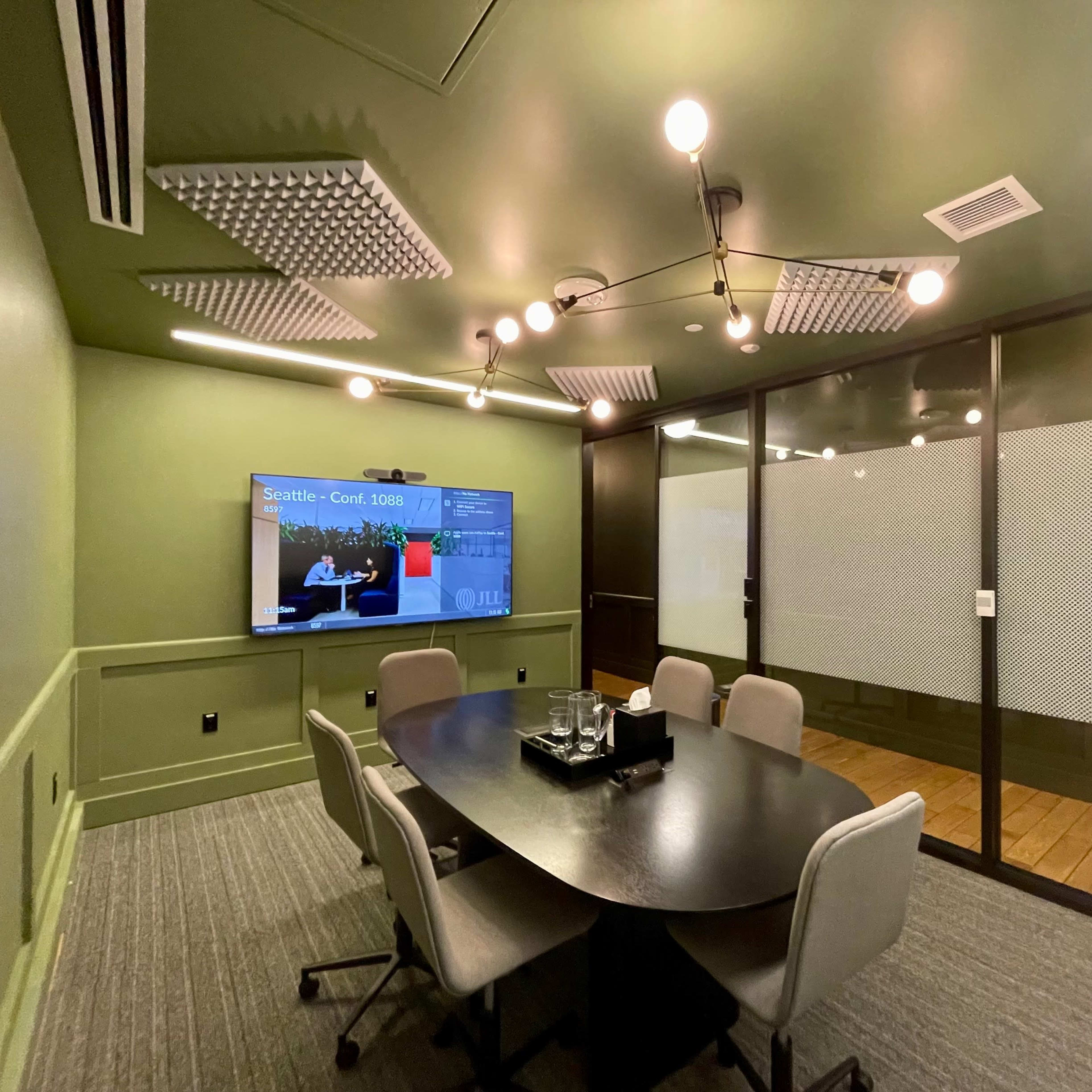 A modern conference room features a round table with chairs, a wall-mounted screen, and a glass-walled side room.