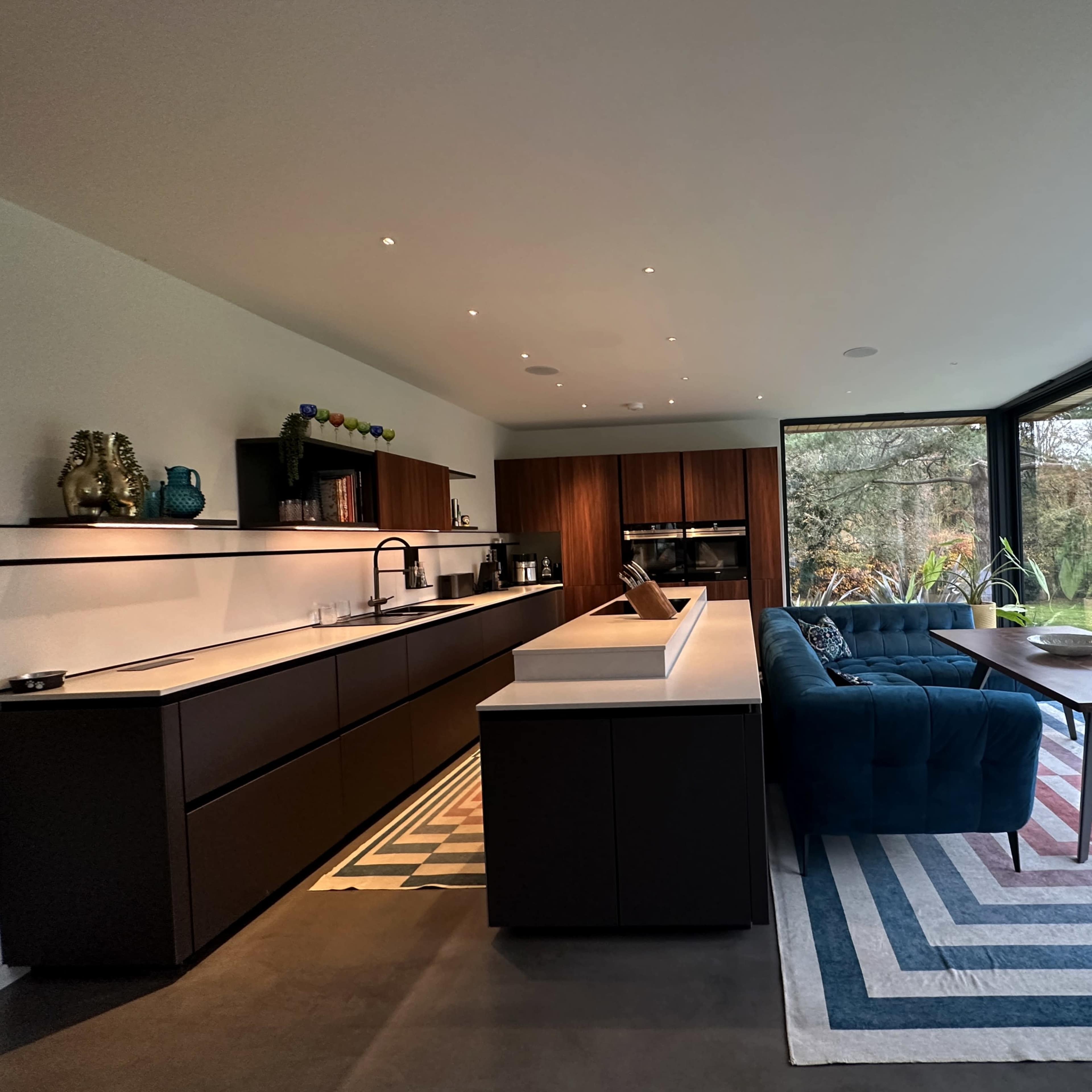 A modern kitchen with dark cabinetry, a white countertop, and a living area featuring a blue sofa and a table, all designed with large windows overlooking a green landscape.
