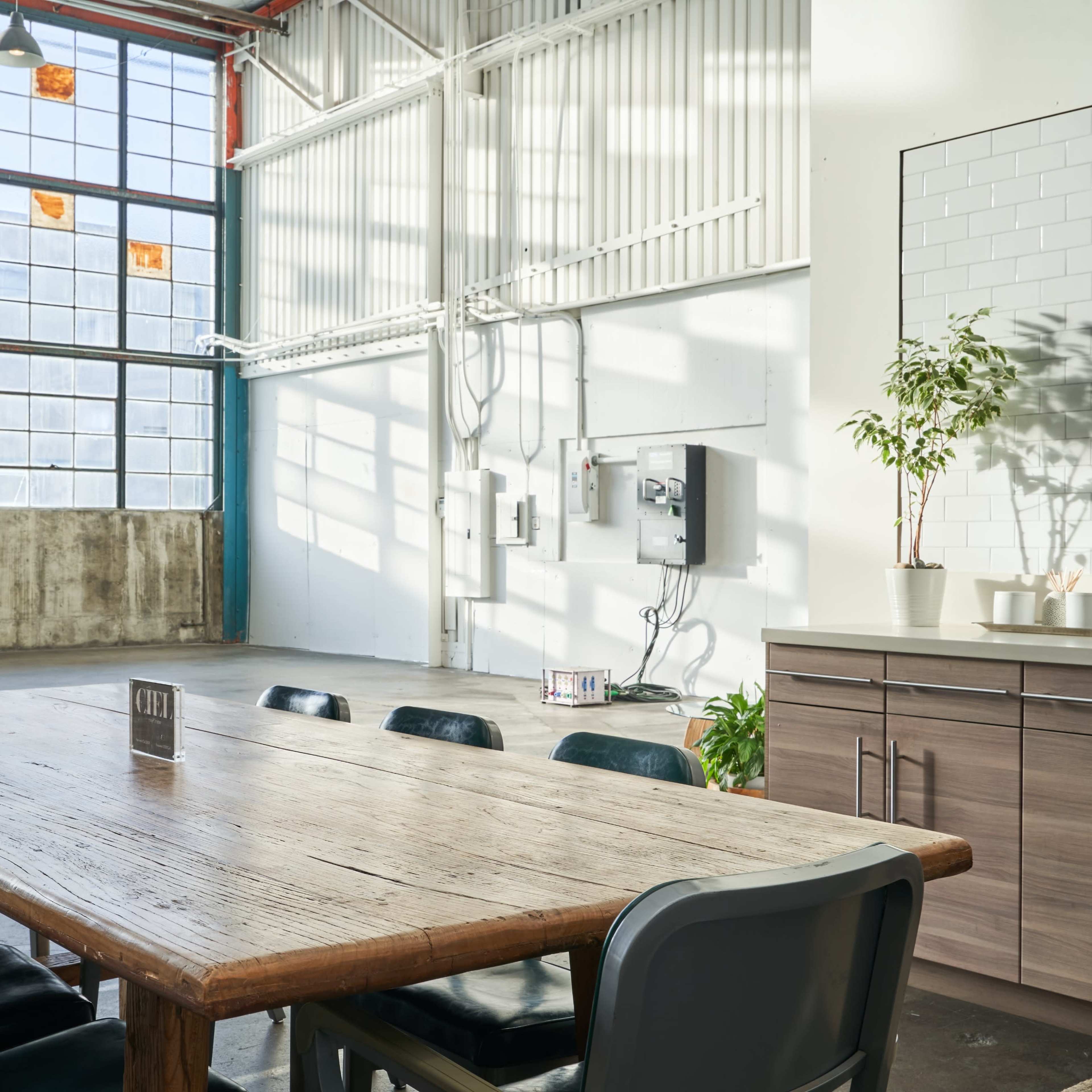 A spacious, well-lit meeting room features a large wooden table surrounded by chairs, with large windows allowing natural light to stream in.