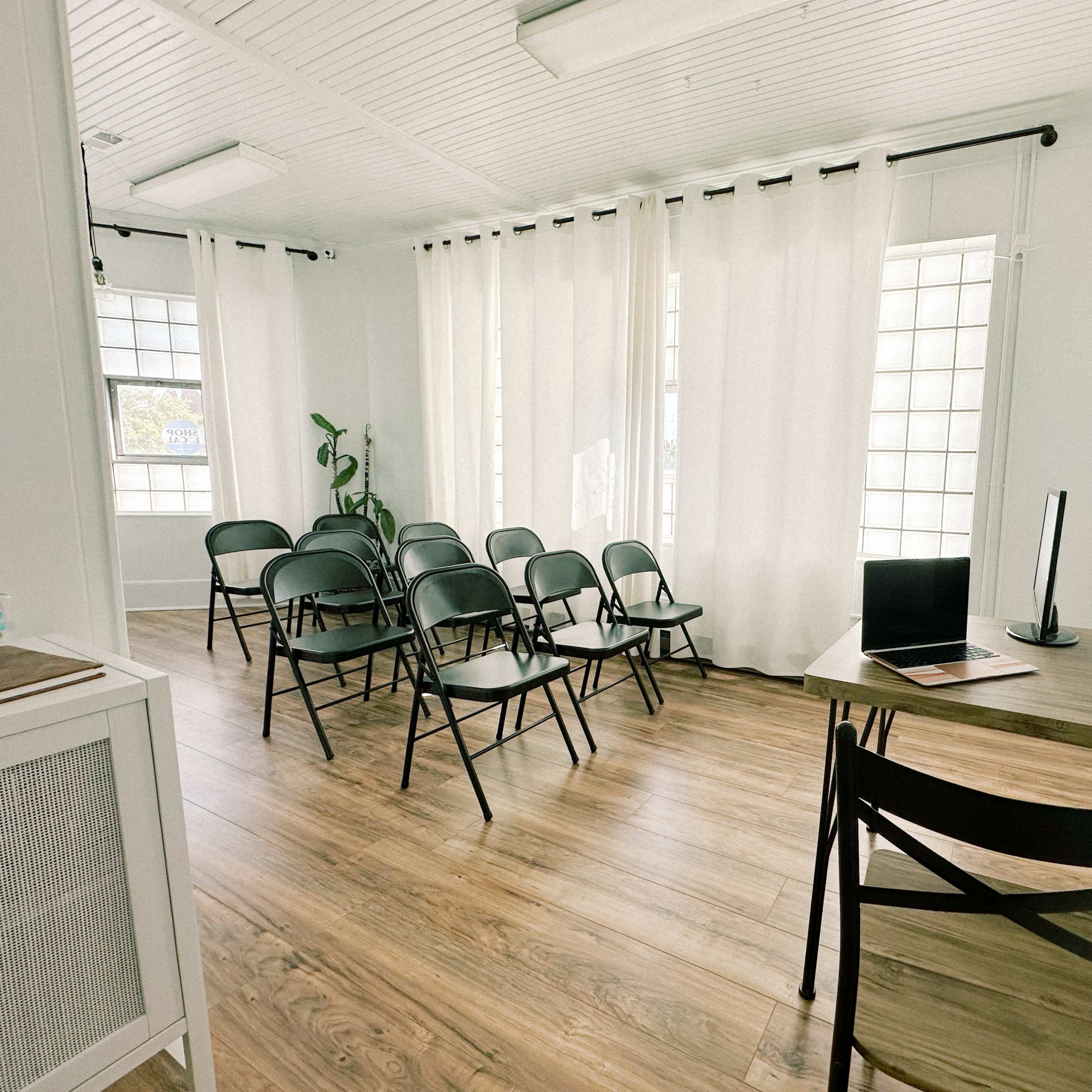 A minimalist meeting room with several black chairs arranged in a semi-circle around a desk with a laptop.