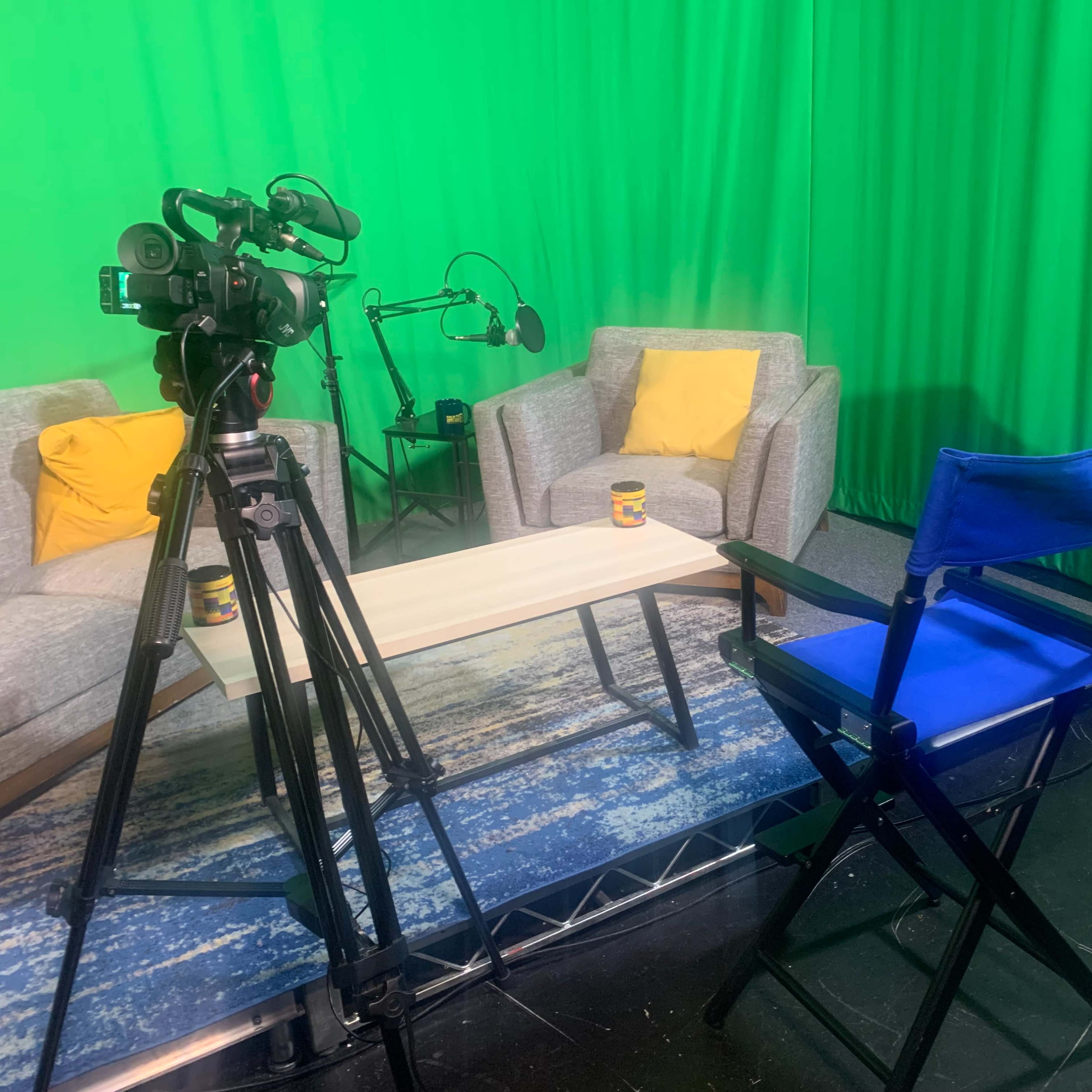 A television studio setup with two gray chairs, a coffee table, and a green backdrop, surrounded by cameras and a microphone.