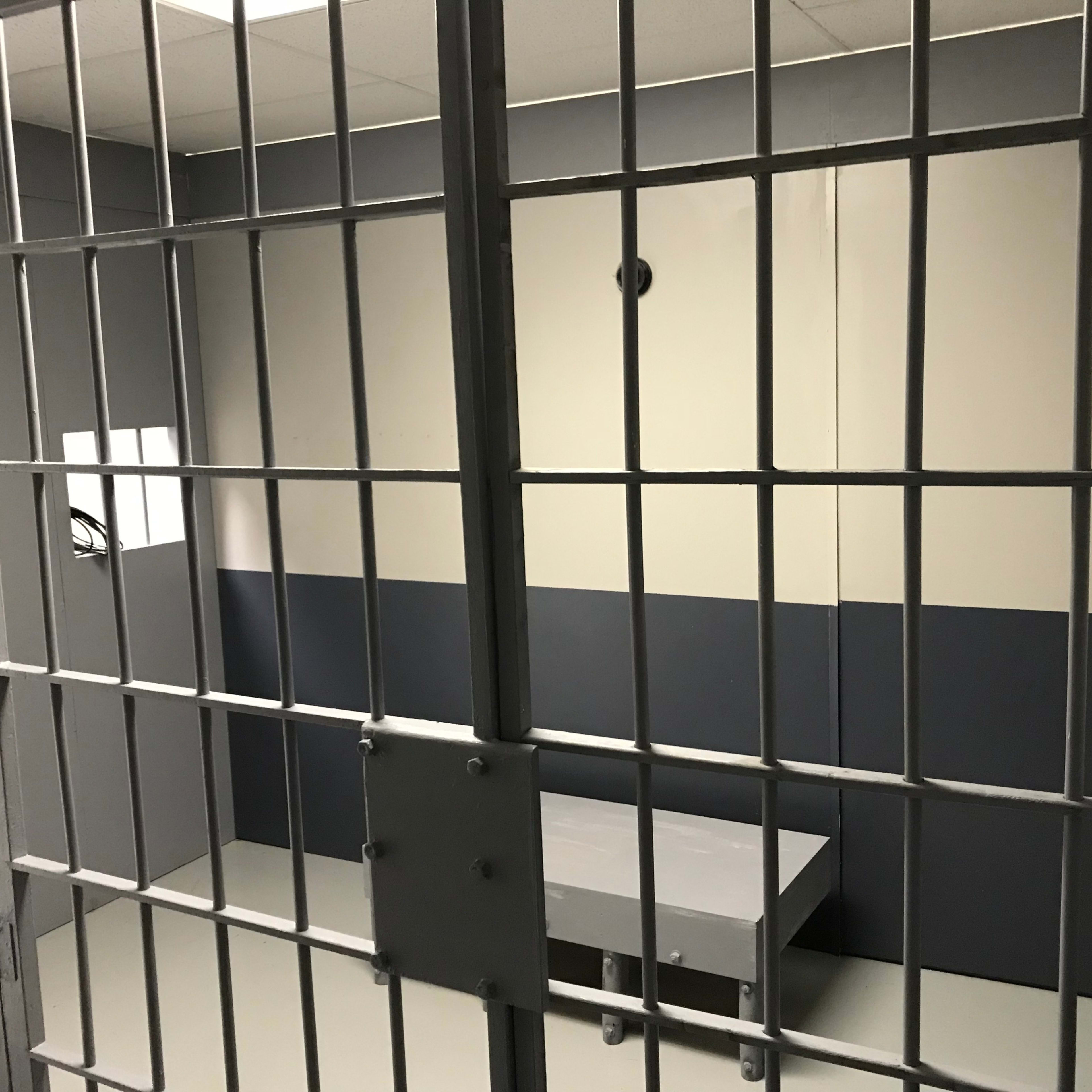The inside of a small, empty jail cell with bars and a plain concrete table.