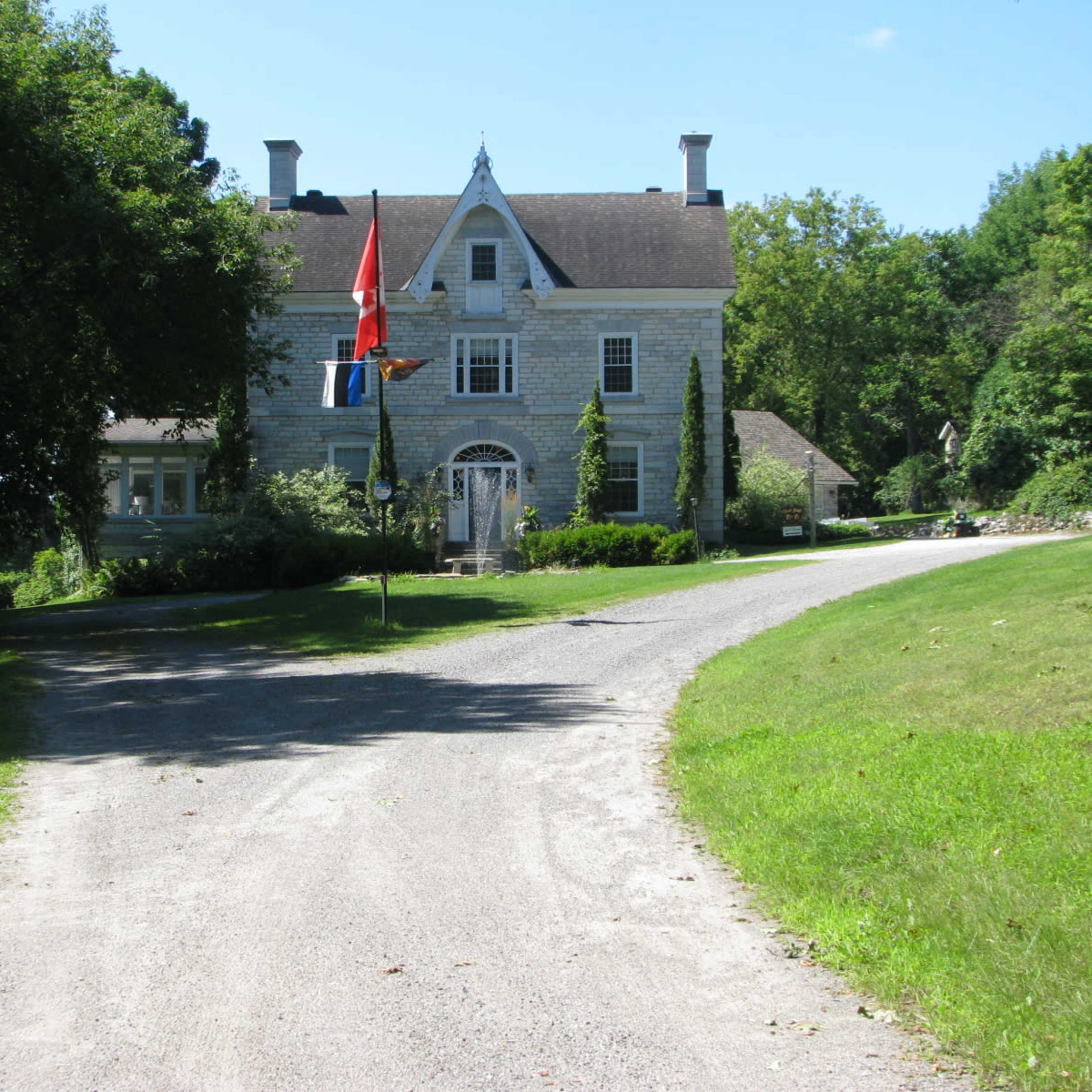 A gravel driveway leads up to a large, historic house surrounded by trees and greenery, with flags displayed in front.