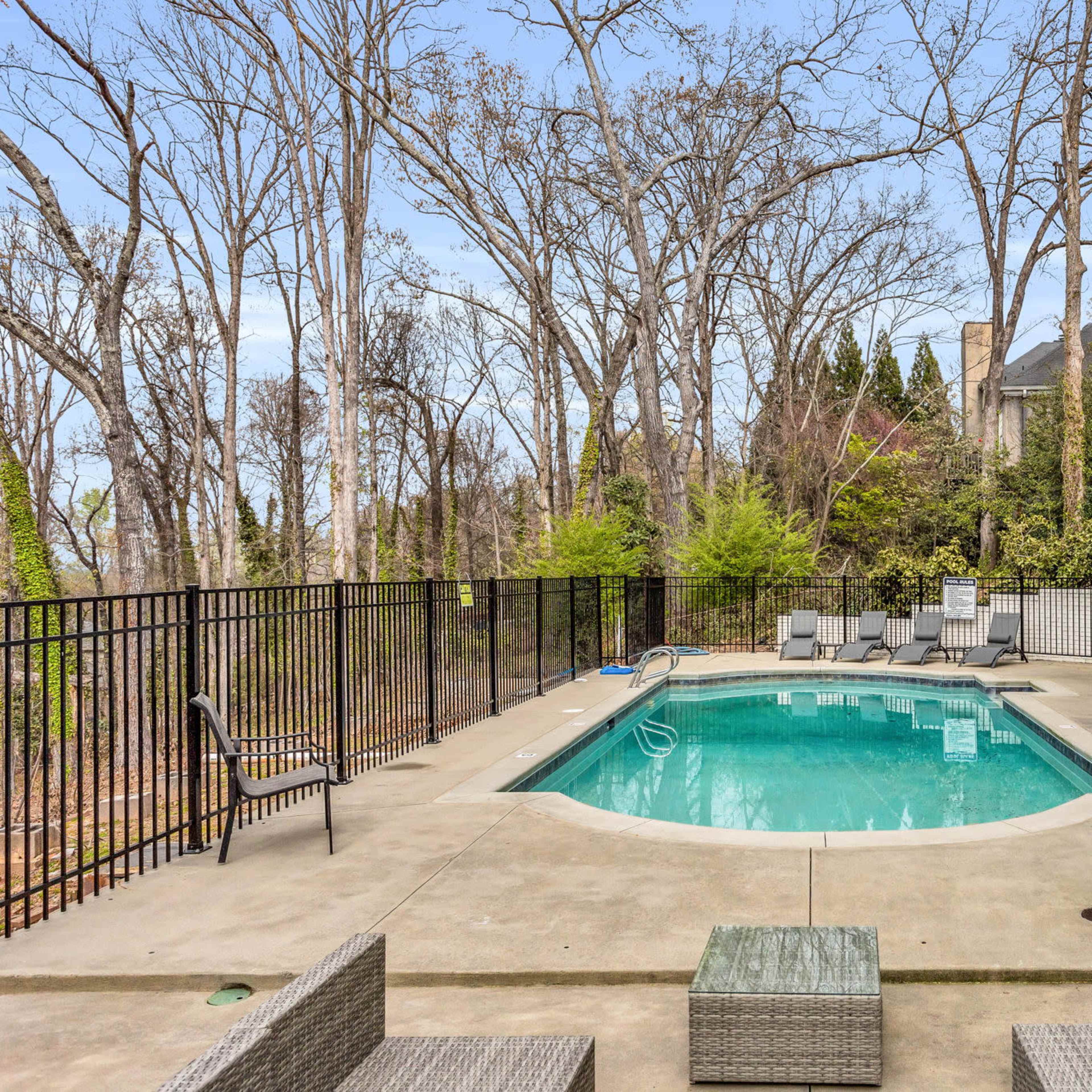 A backyard pool area surrounded by tall trees, with lounge chairs and a fence enclosing the pool.