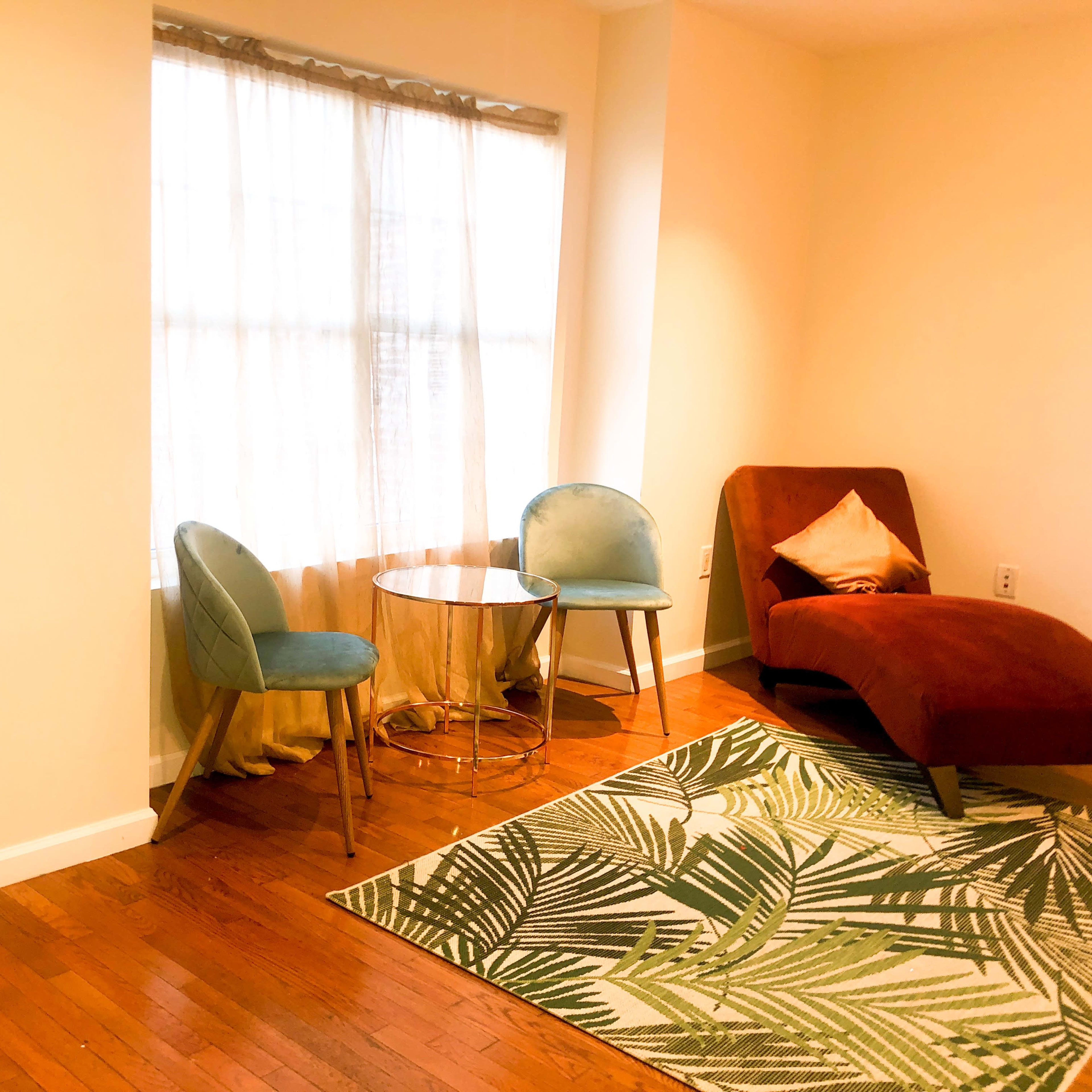 A modern living space with two chairs, a small round table, and a chaise lounge, illuminated by natural light coming through a window, with a patterned rug on the wooden floor.