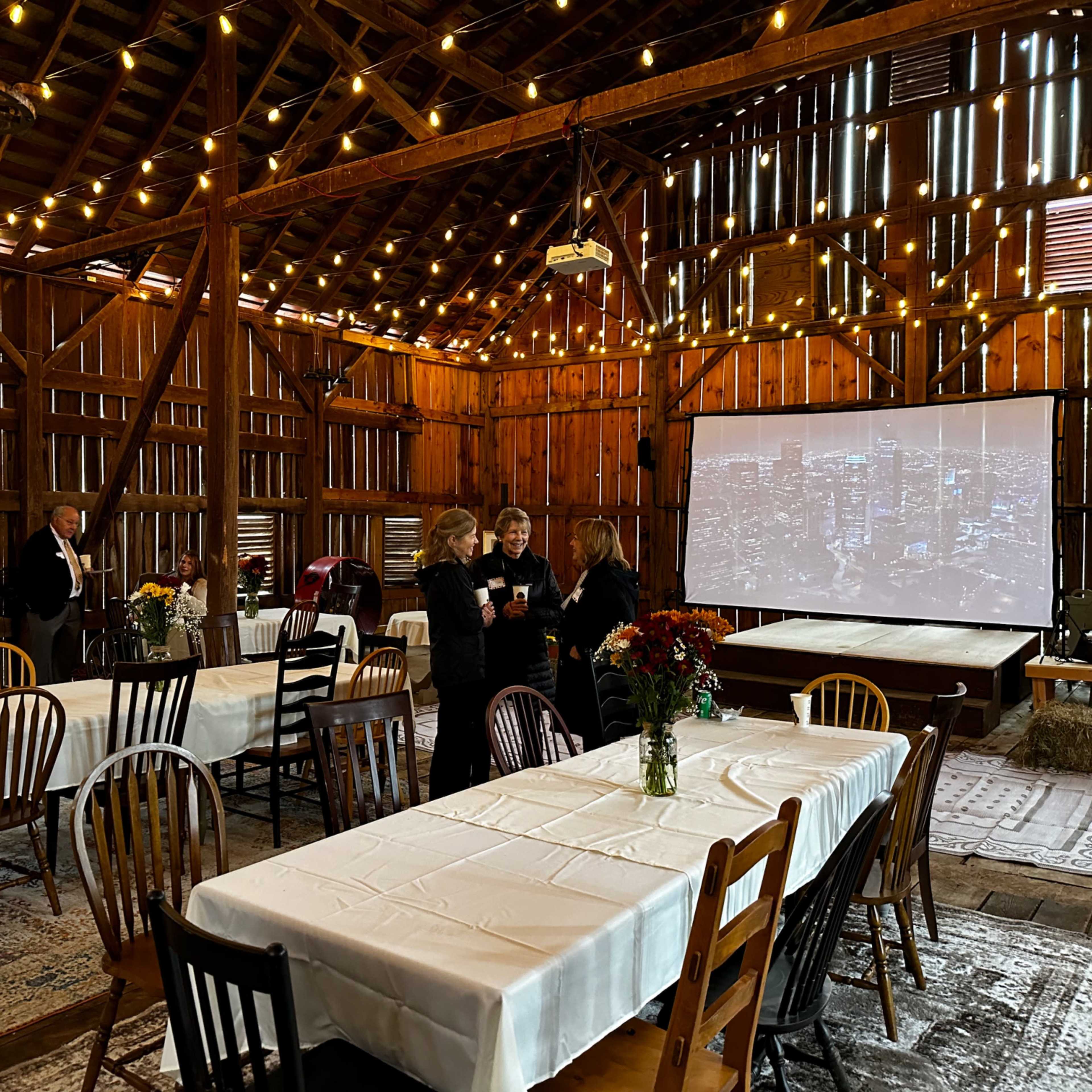 A rustic barn interior is set up for an event, featuring tables with white tablecloths, wooden chairs, and string lights overhead, while guests converse near a projection screen showing a city skyline.