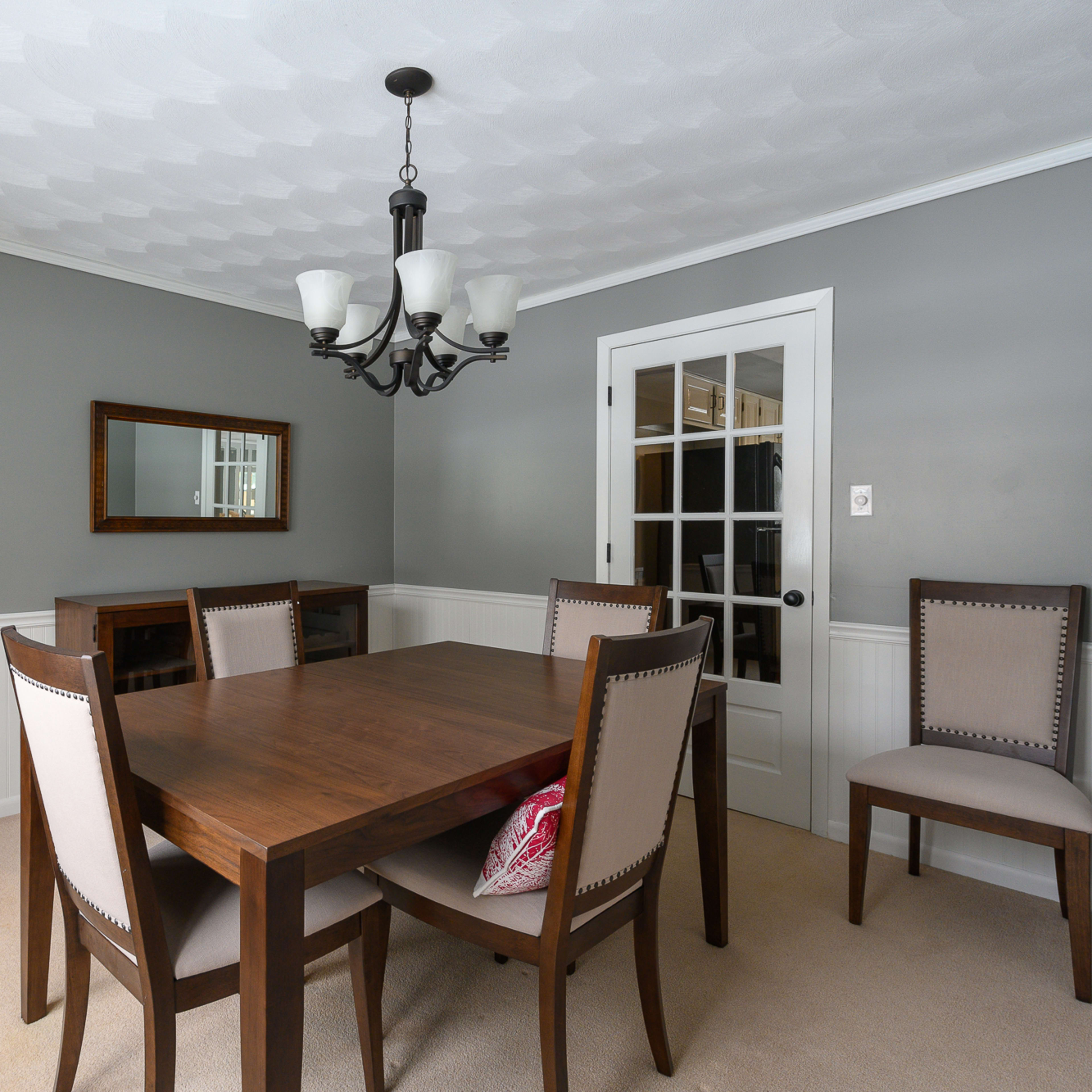 A dining room features a wooden table surrounded by upholstered chairs, with a chandelier hanging above and a mirror on the wall.