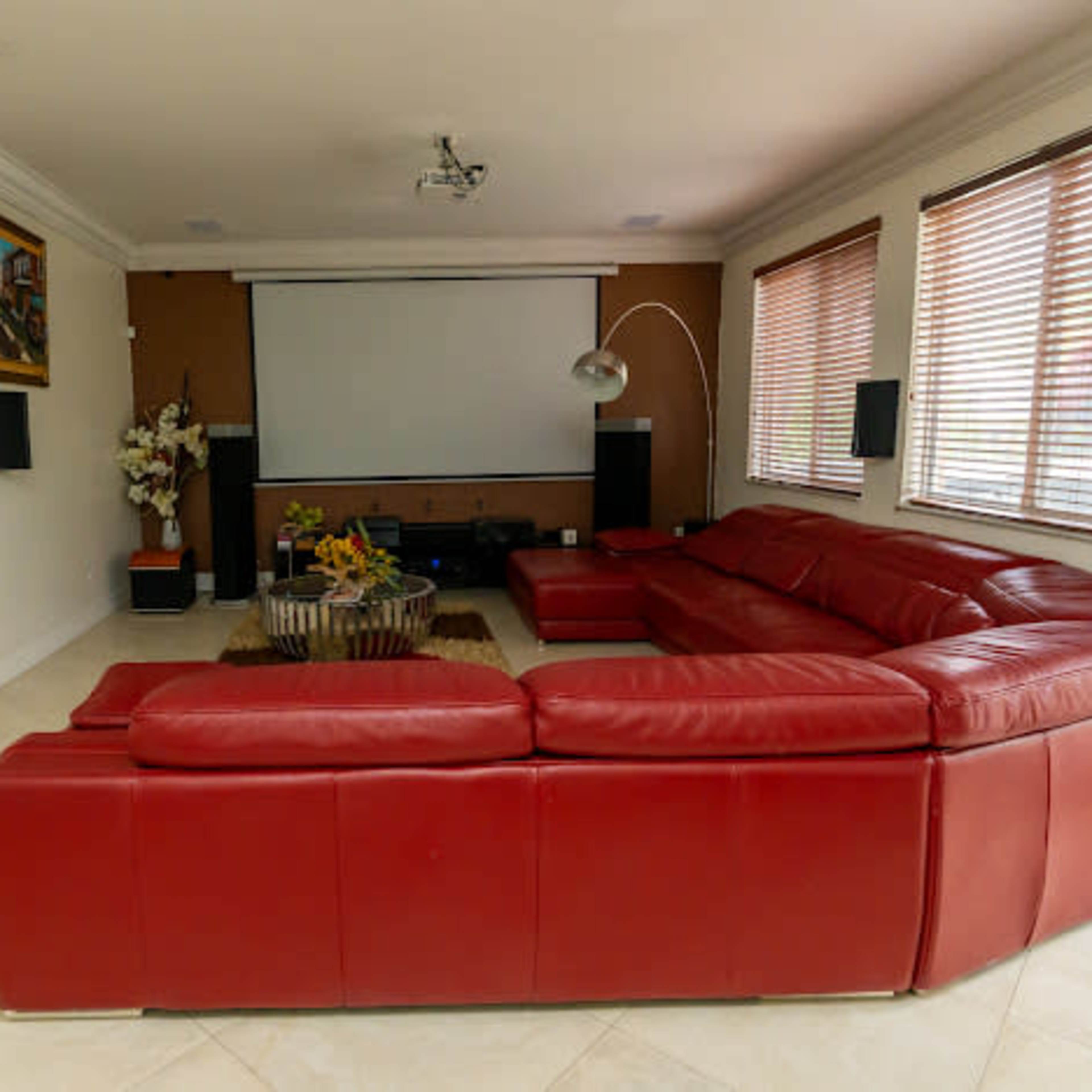A modern living room features a large red sectional sofa, a wall-mounted projector screen, and natural light filtering through window blinds.