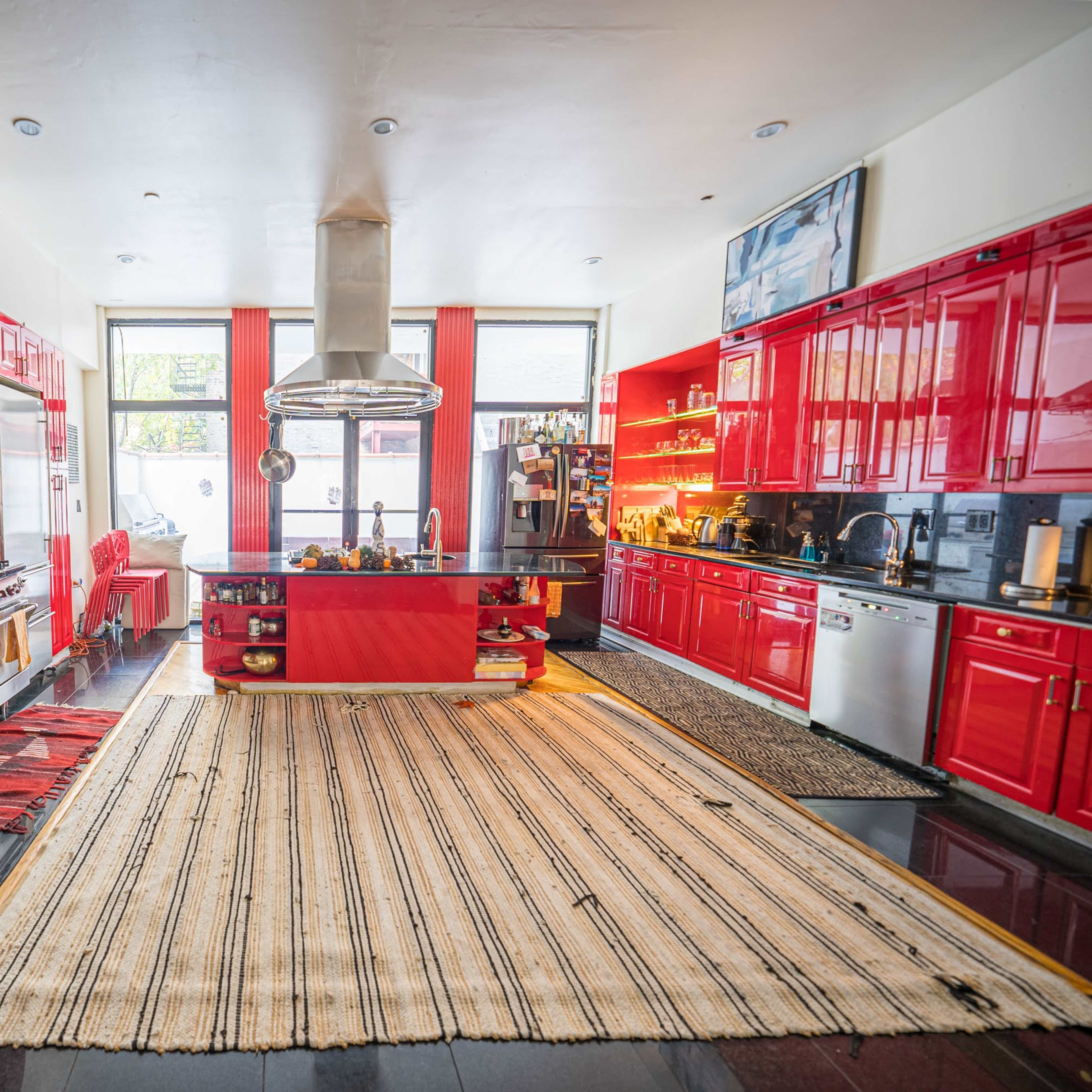 A spacious kitchen features red cabinetry, stainless steel appliances, and a central island with a polished surface, surrounded by patterned rugs.