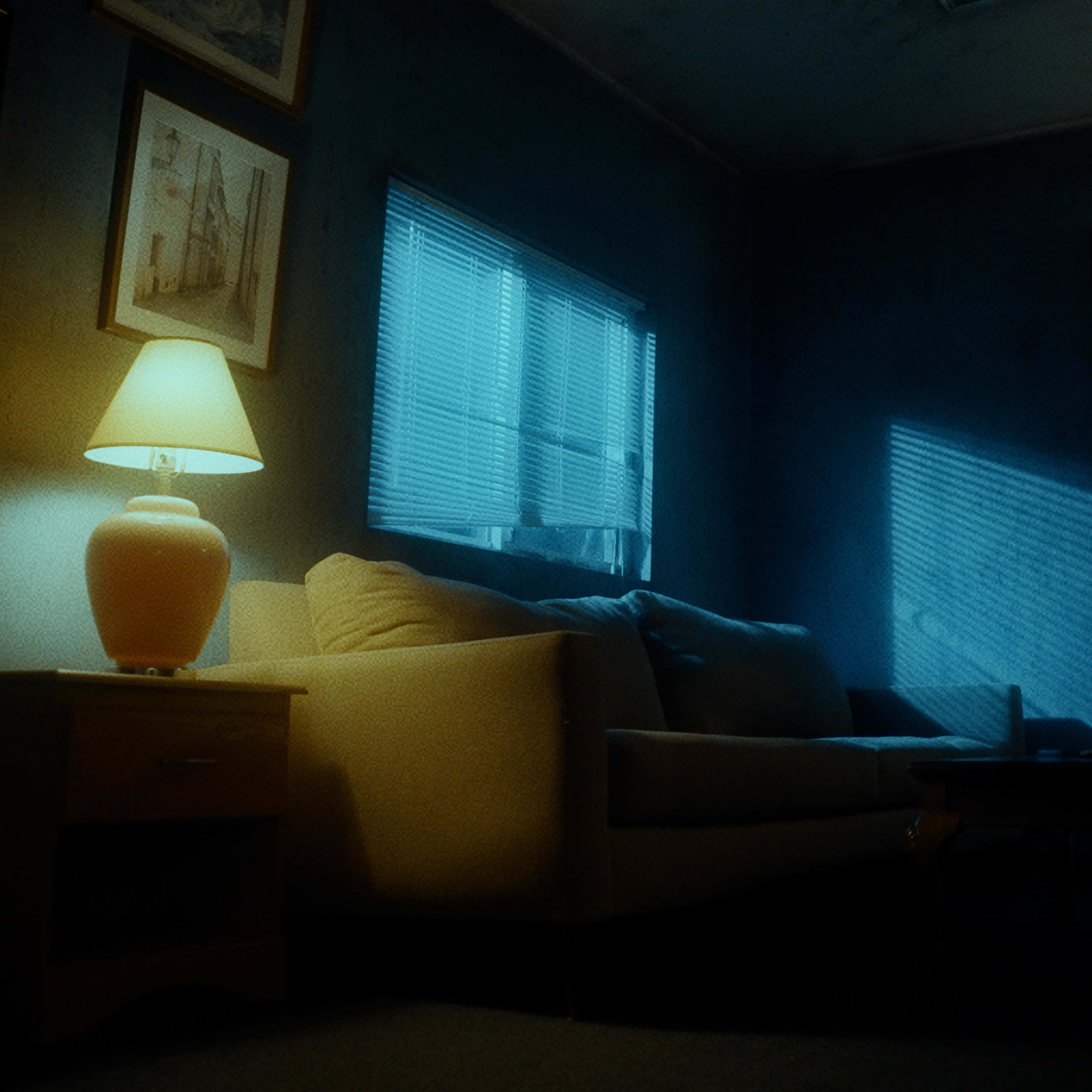 A dimly lit living room features a beige couch, a coffee table, and a lamp casting soft light, with shadows from the window blinds creating patterns on the floor.