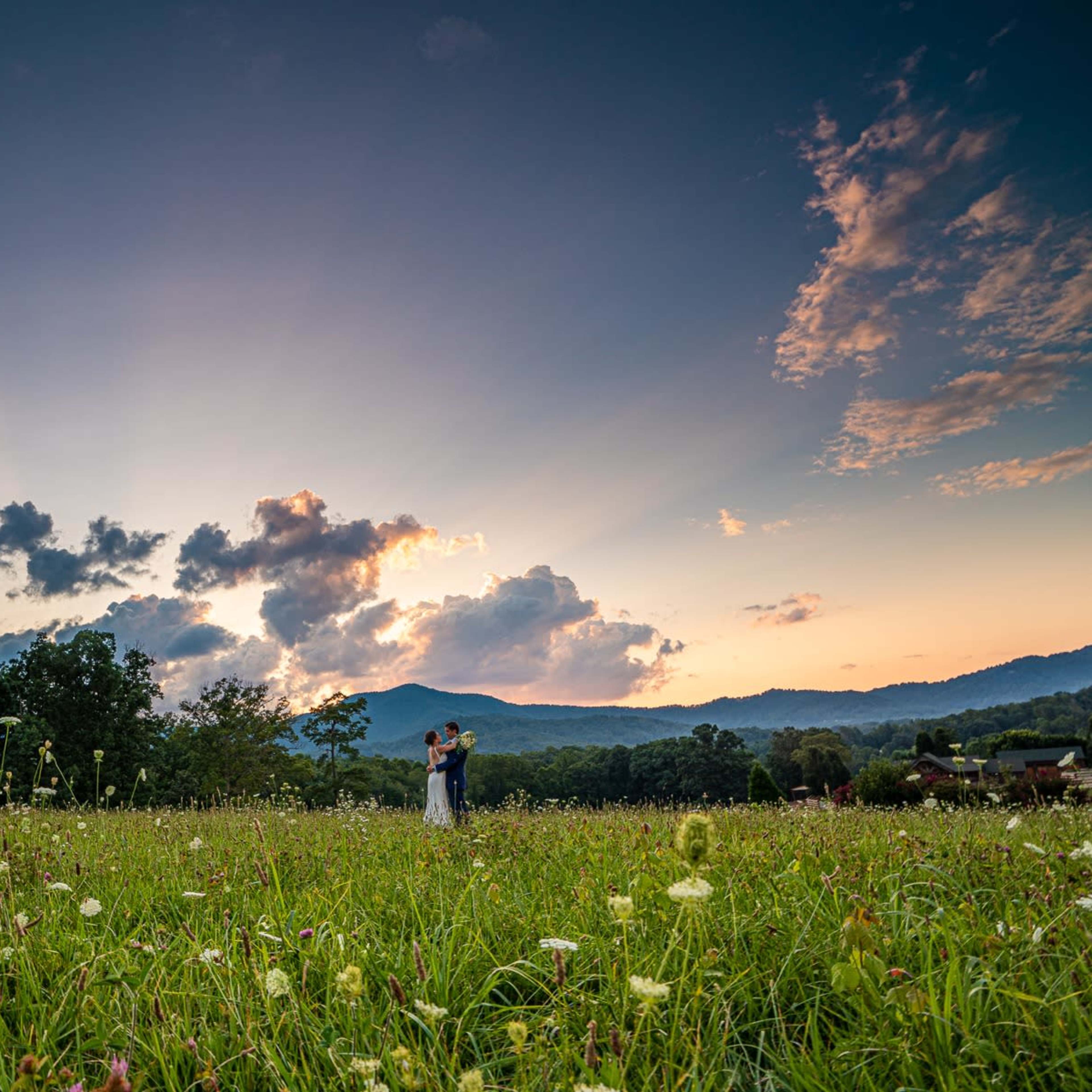 A couple embraces in a grassy field under a sky filled with clouds and rays of sunlight at sunset.