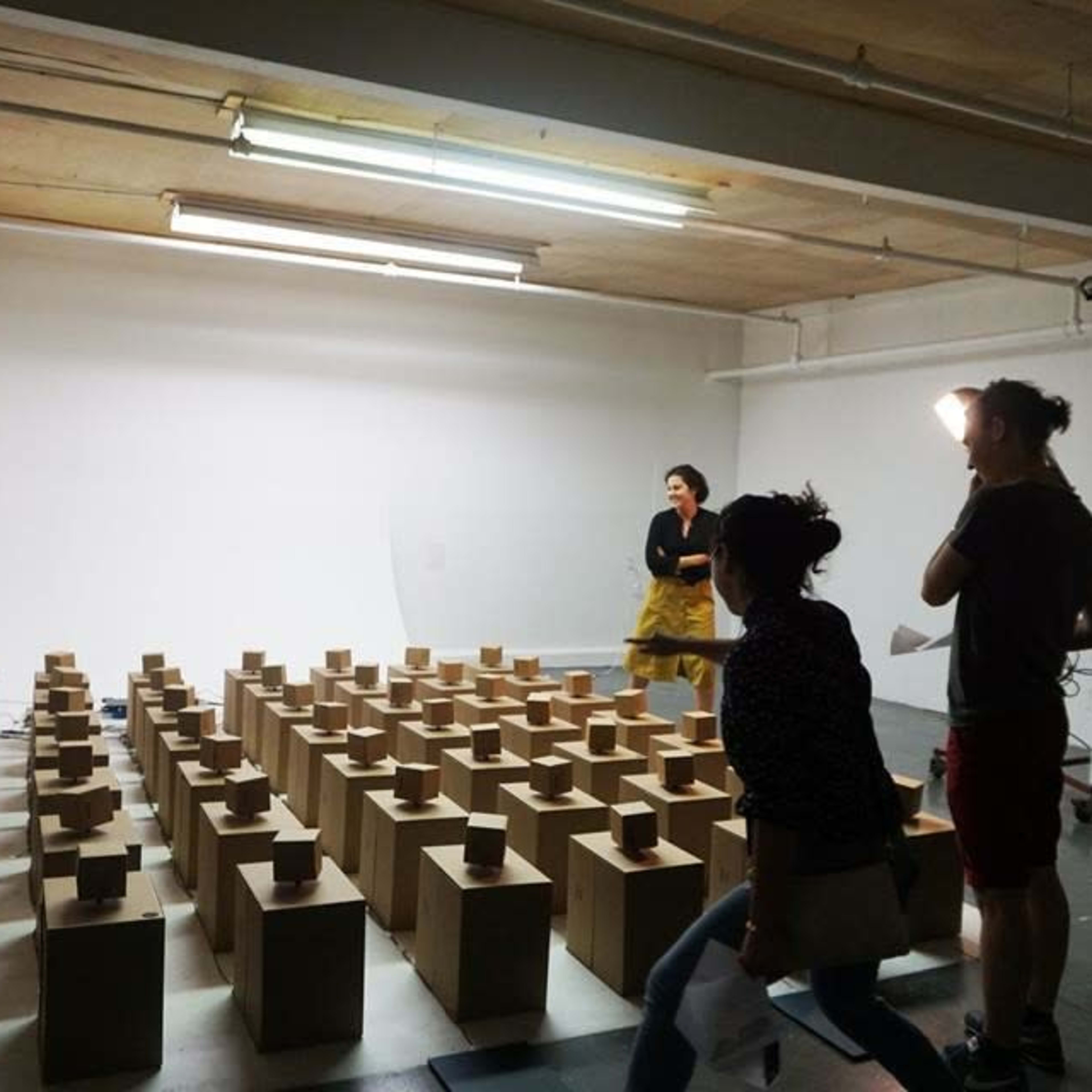 A studio space is organized with numerous wooden boxes arranged in rows, while two people are positioned in the background.