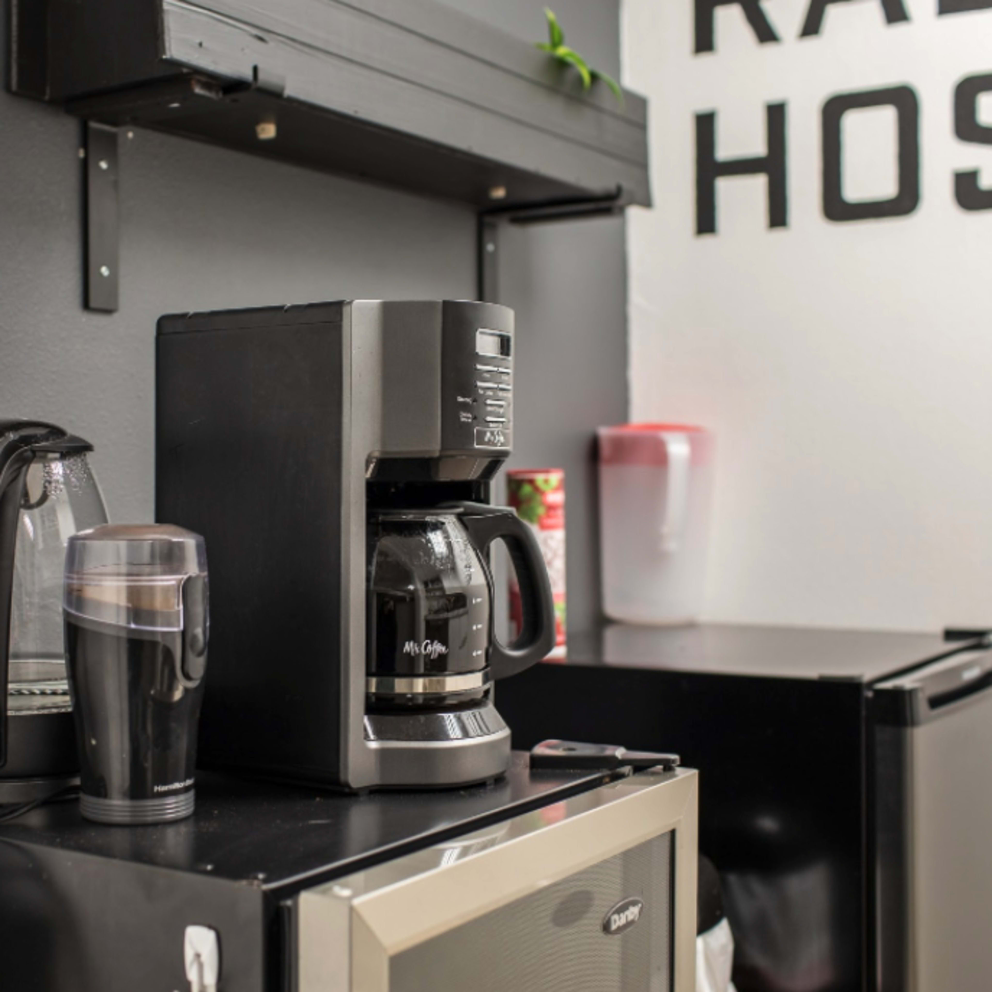 A coffee maker is placed on a countertop next to a stainless steel travel mug and a small refrigerator in a kitchen setting.