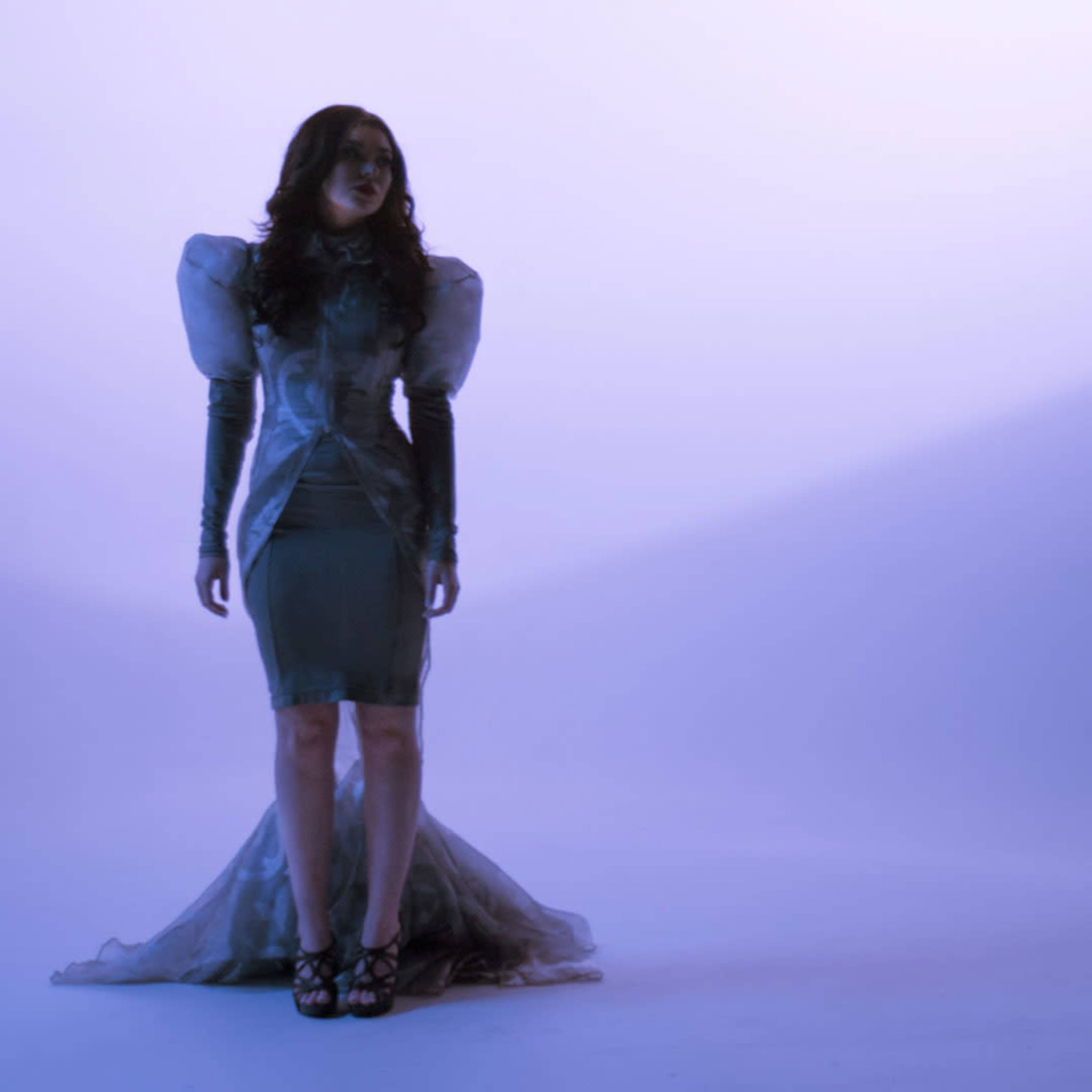 A person stands in a fitted dress with dramatic shoulder details against a soft purple backdrop.