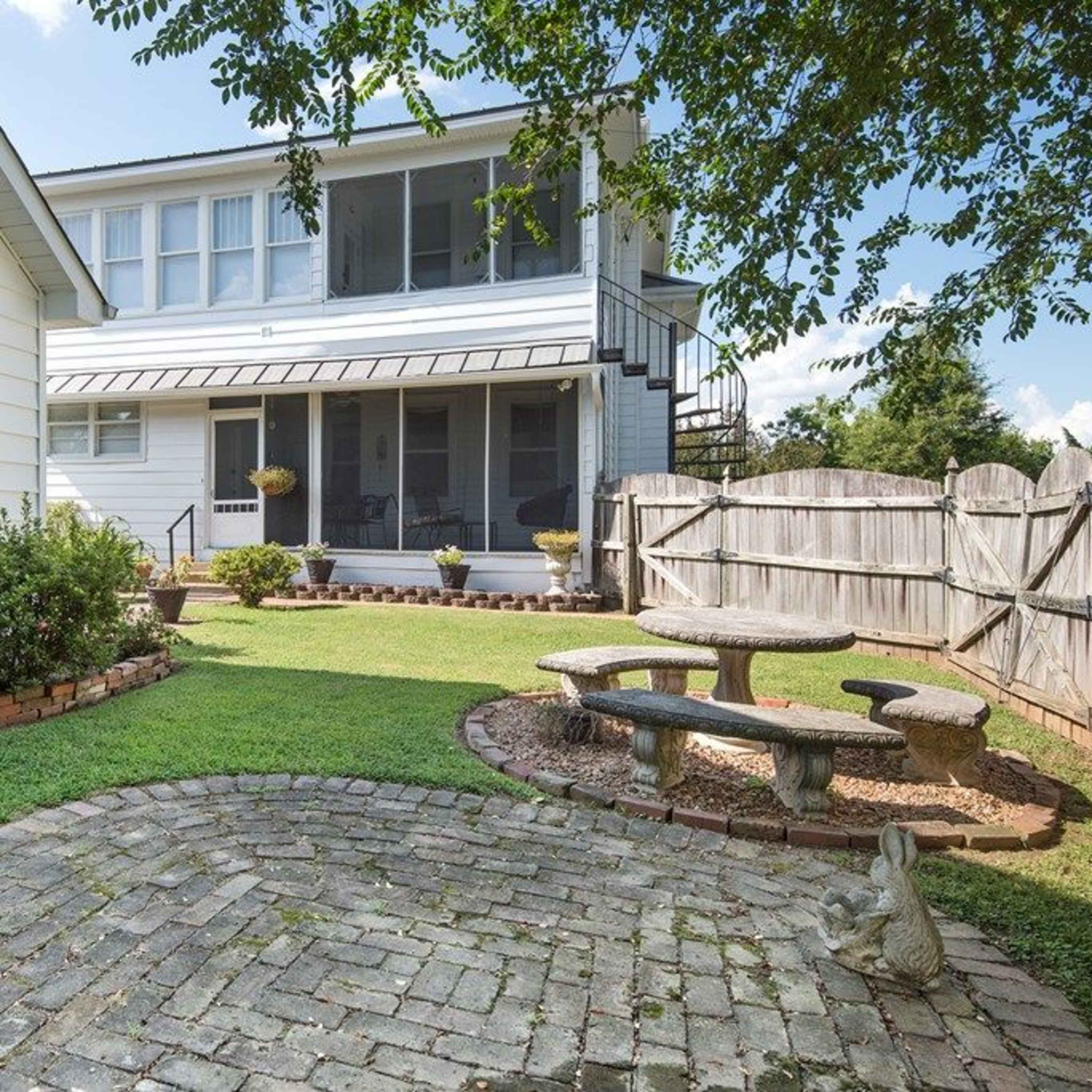 A backyard with a stone patio, bench seating, and a grassy area surrounded by a wooden fence and a two-story house.