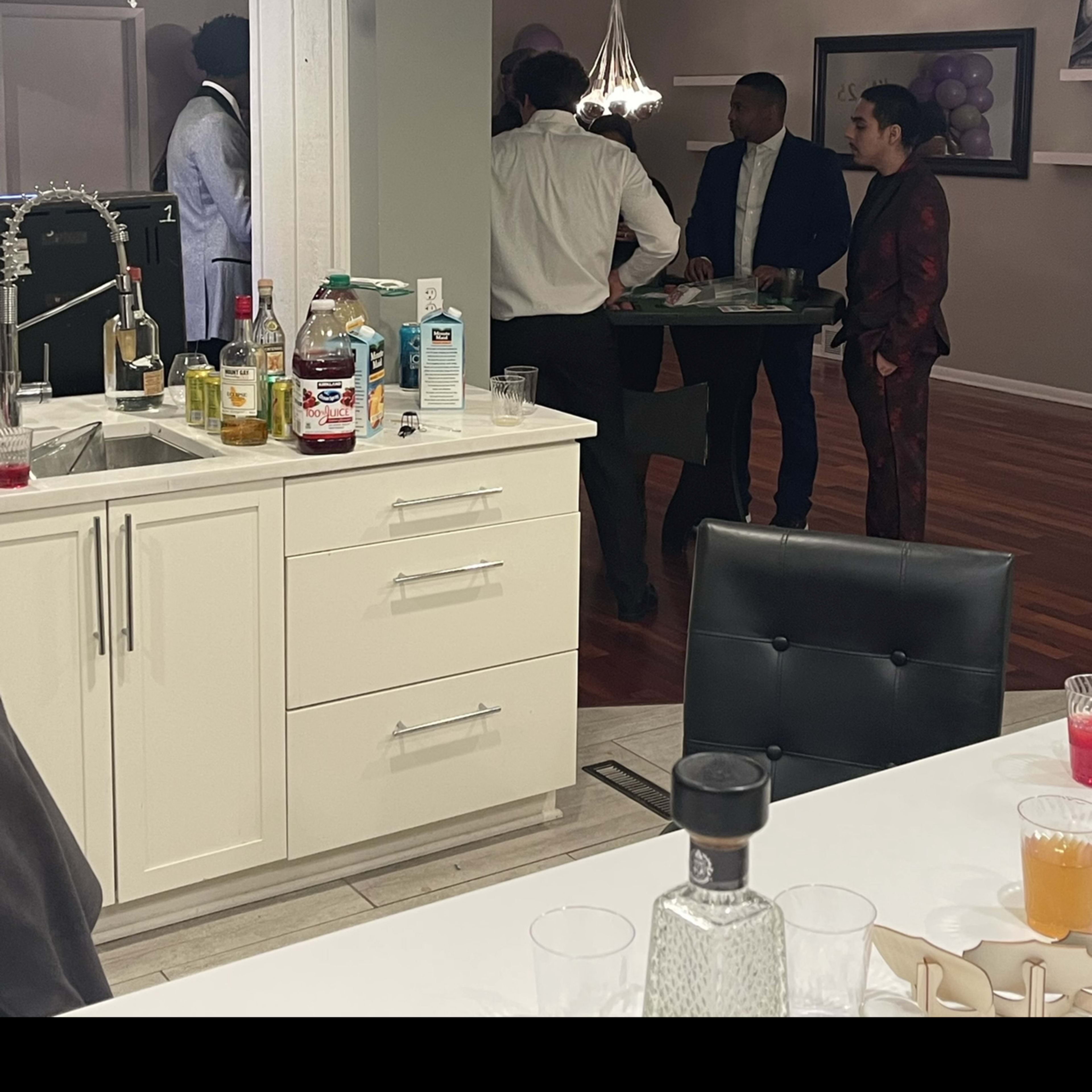 A group of people dressed in formal attire stands around a table in a well-lit kitchen area.