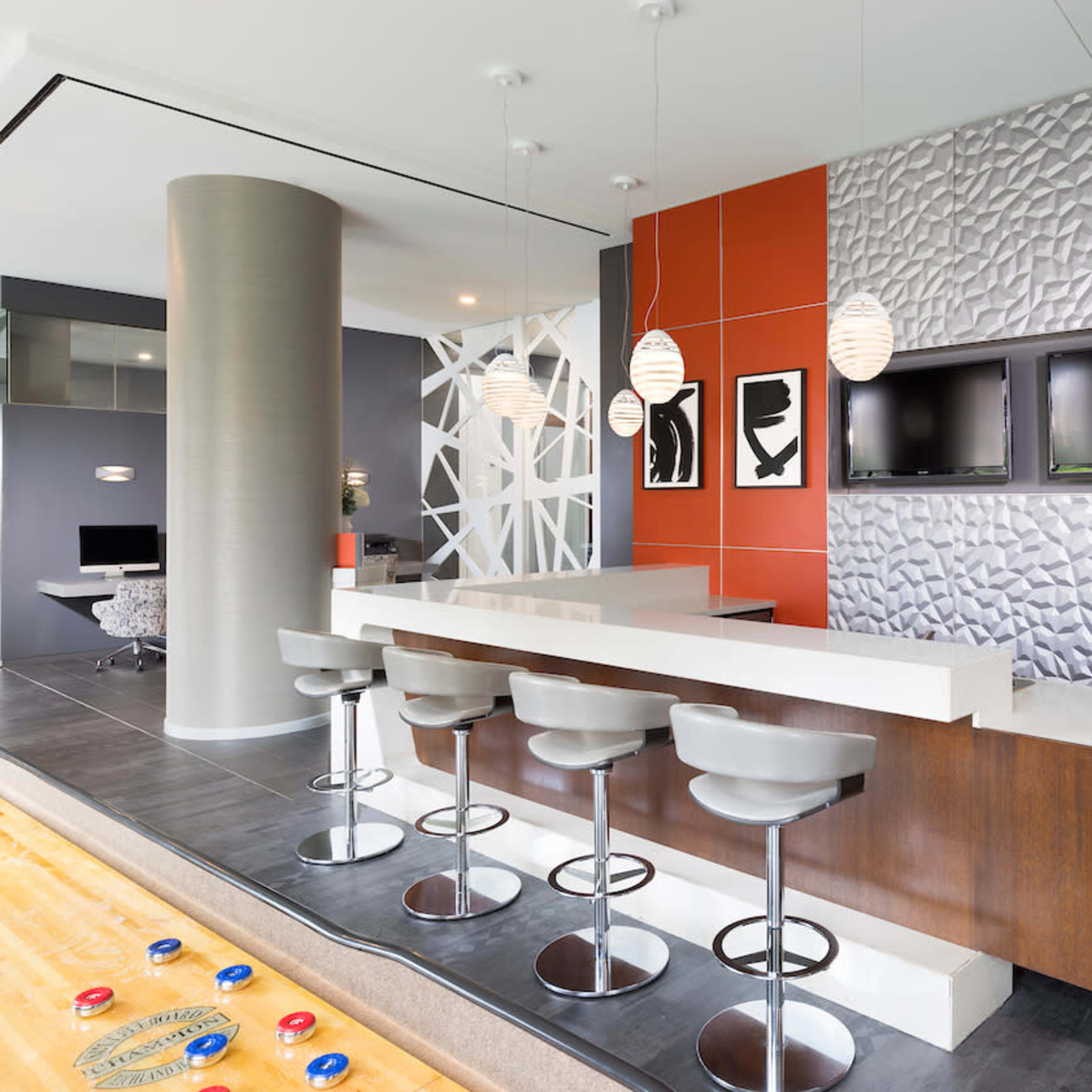 A modern kitchen with a bar area featuring three stools, a textured wall, and two television screens mounted above the countertop.
