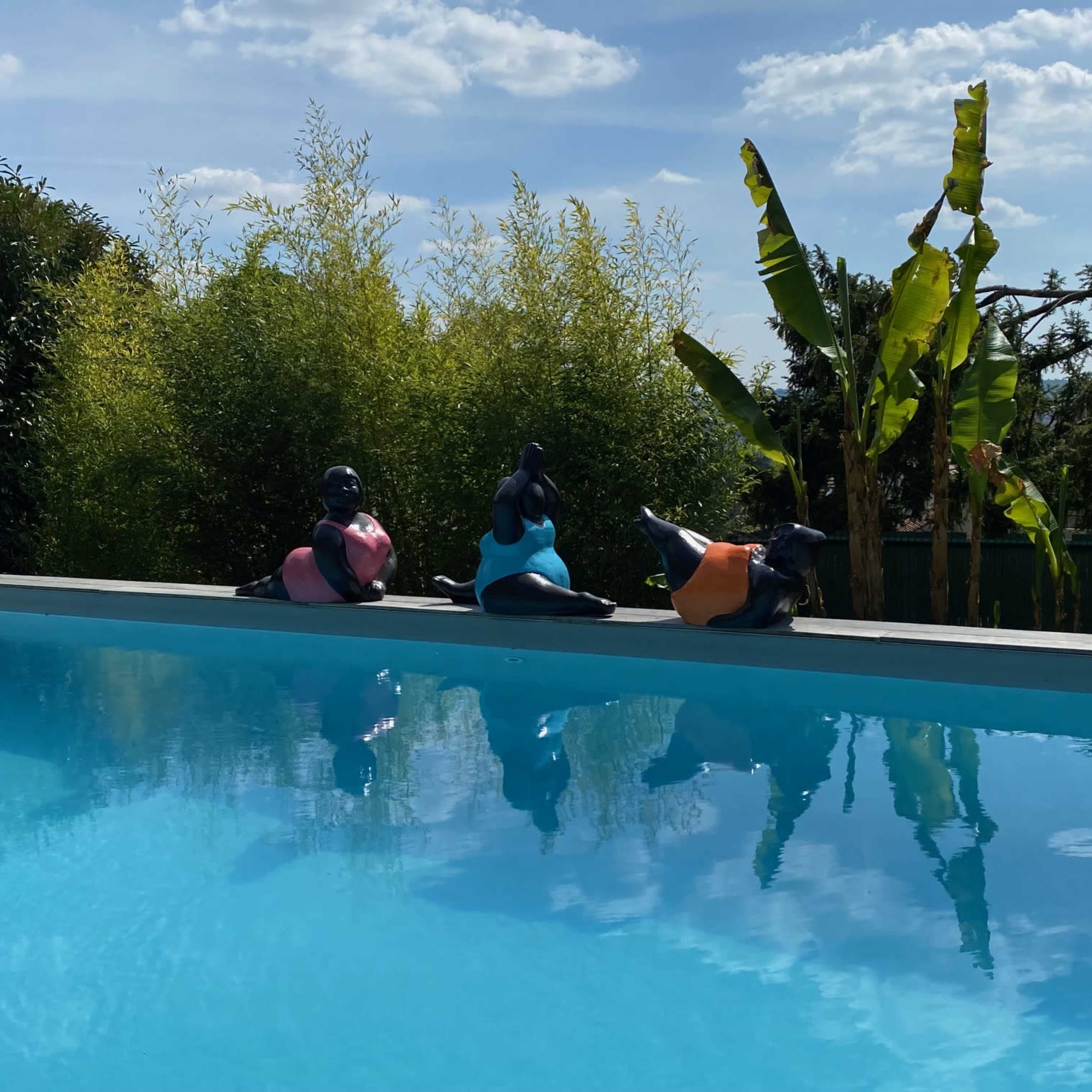 Three colorful sculptures of figures sit on the edge of a swimming pool, reflecting in the water under a blue sky with scattered clouds.