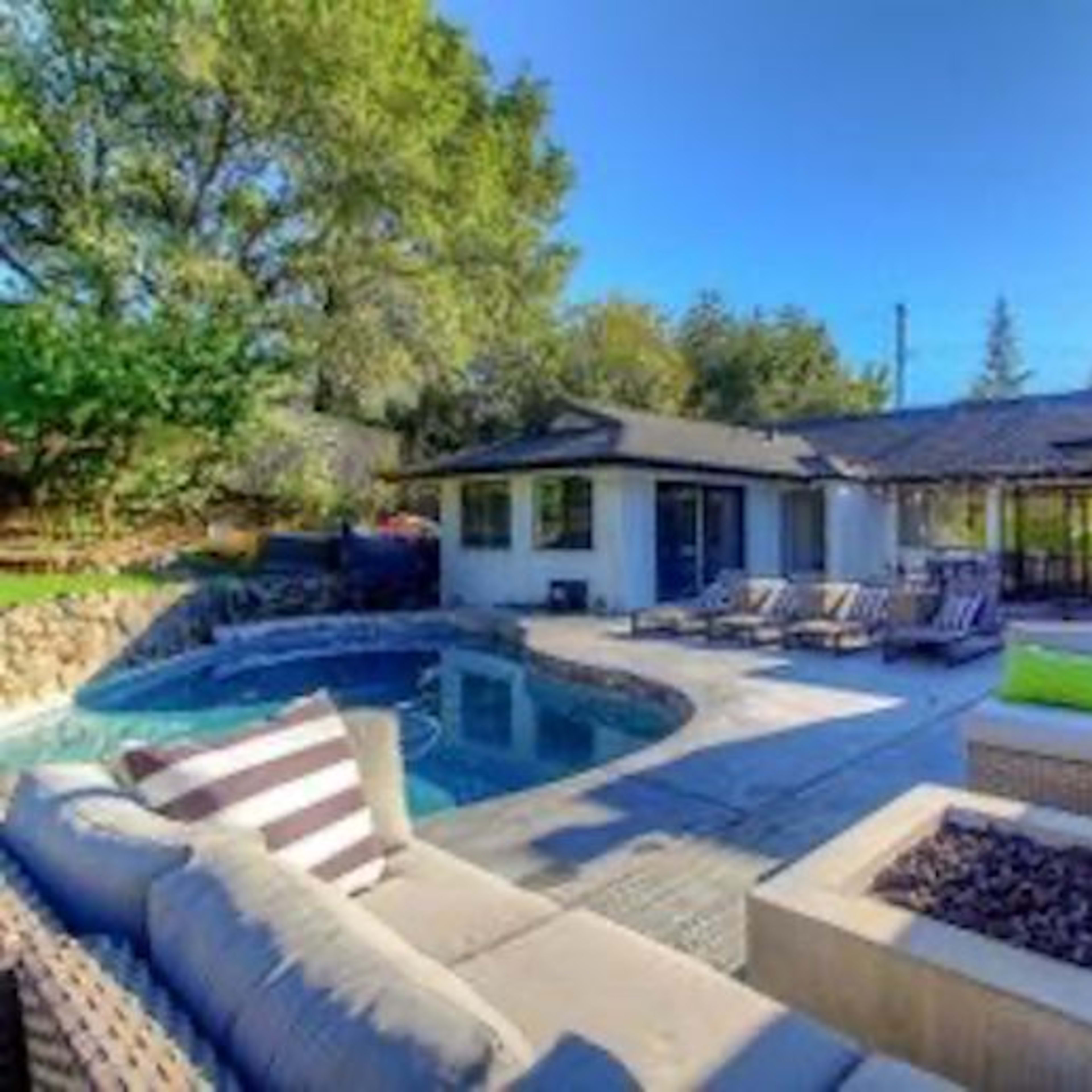 A spacious backyard features a swimming pool surrounded by lounge chairs and a stone patio, with a modern home and landscaped greenery in the background.