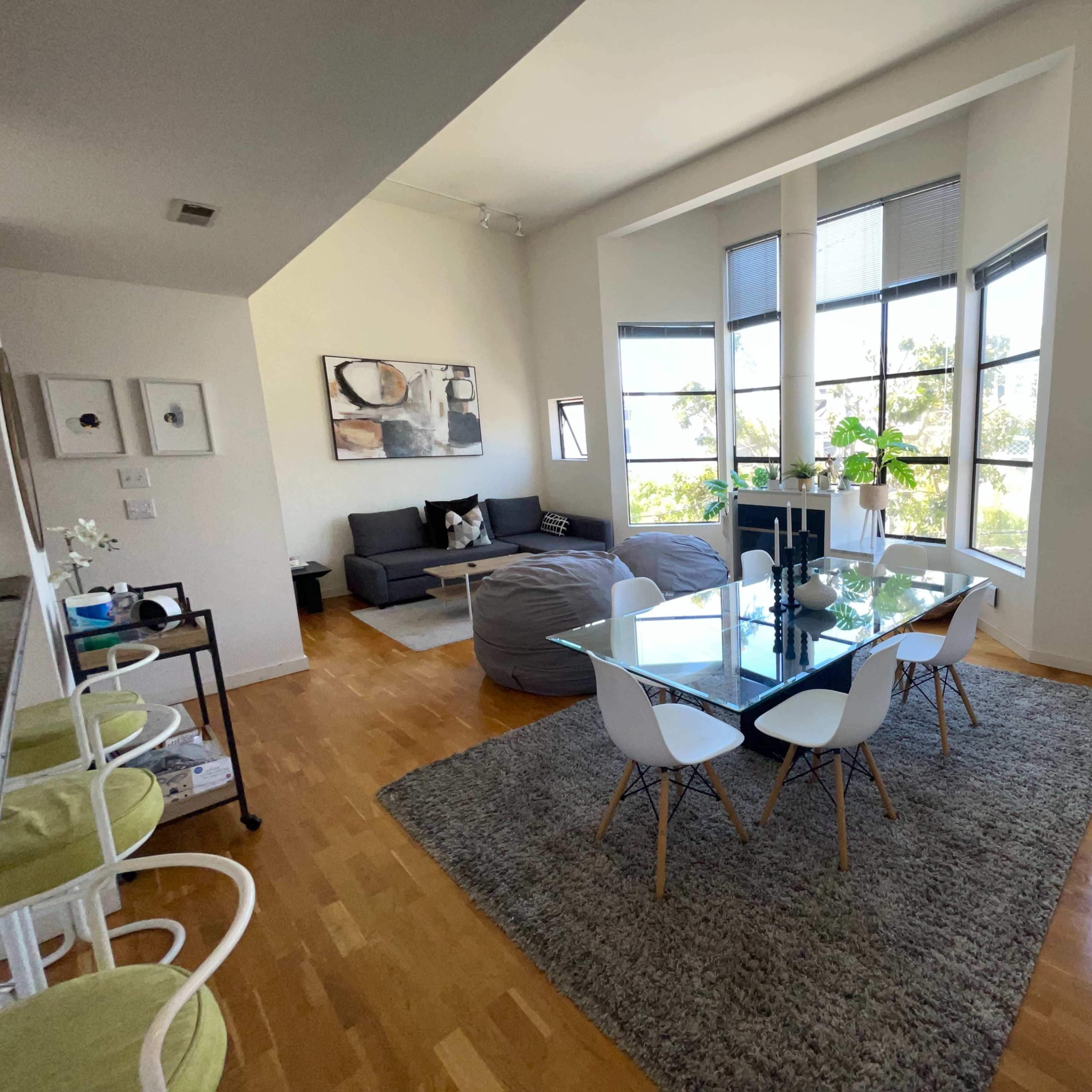 A modern living space featuring a dining area with a glass table and white chairs, a gray sectional couch, and large windows allowing natural light.