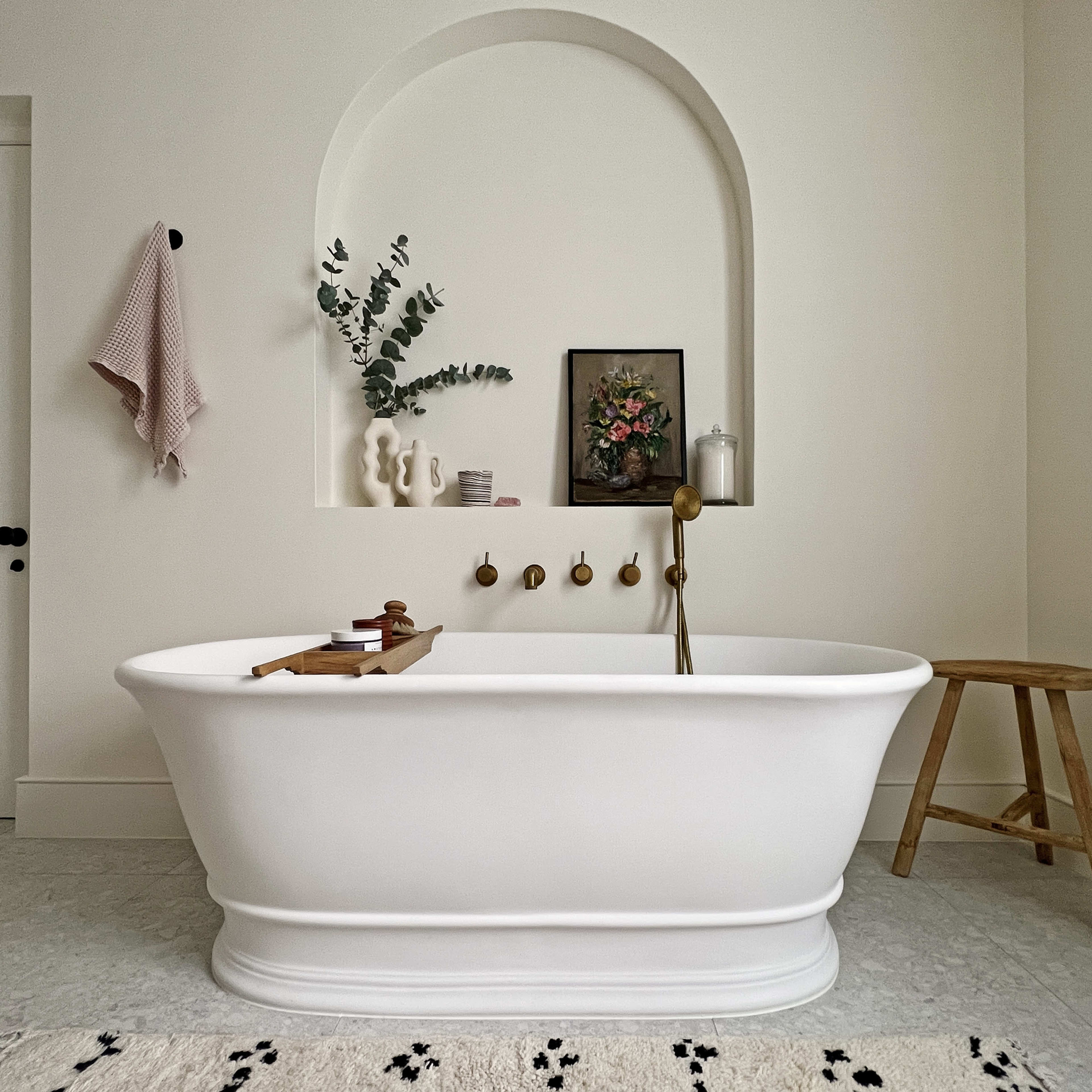 A white freestanding bathtub in a minimalistic bathroom with a textured wall, a wooden stool, and decorative items on a shelf.