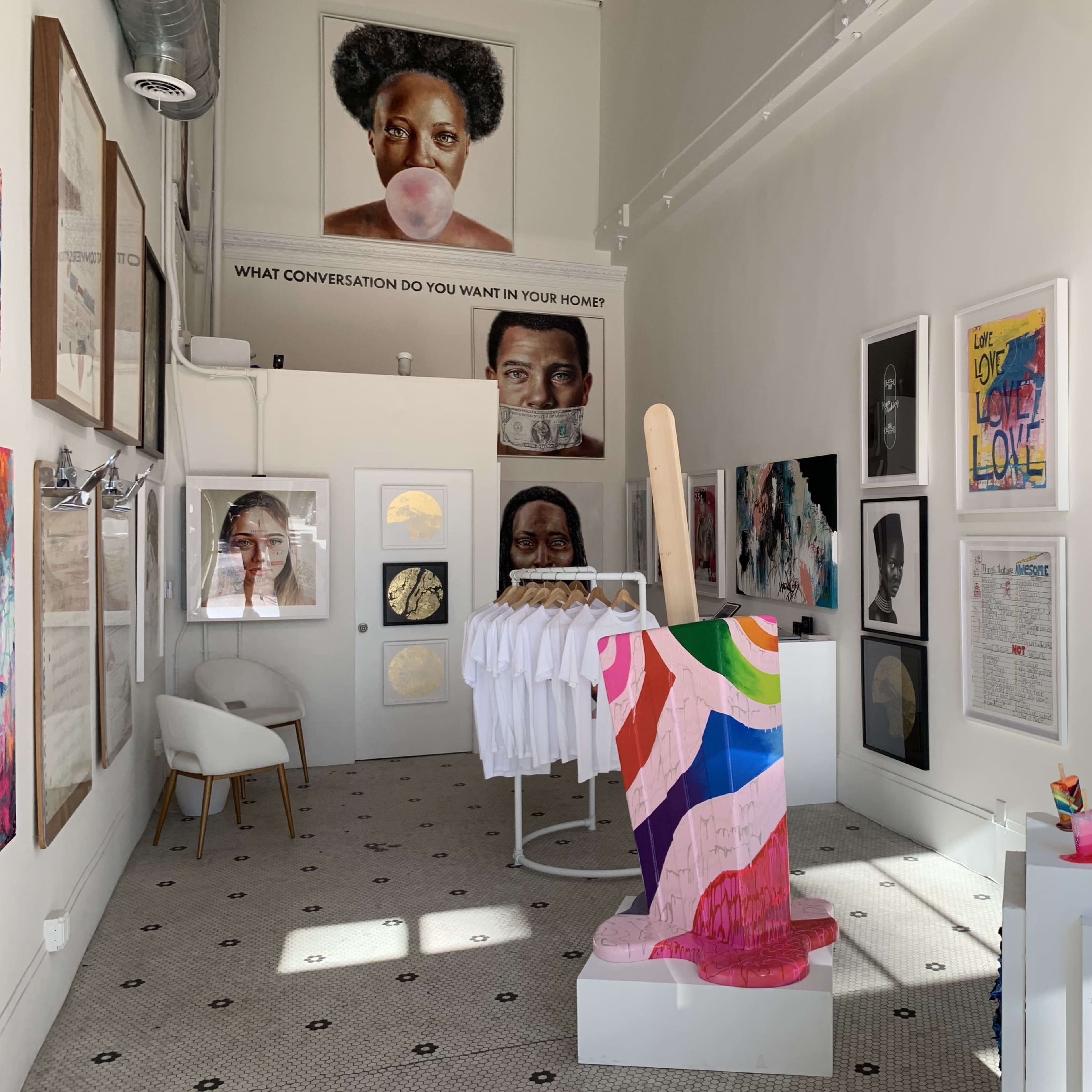 An art gallery featuring various artworks on the walls, including portraits and colorful installations, along with a display of white T-shirts on a rack.