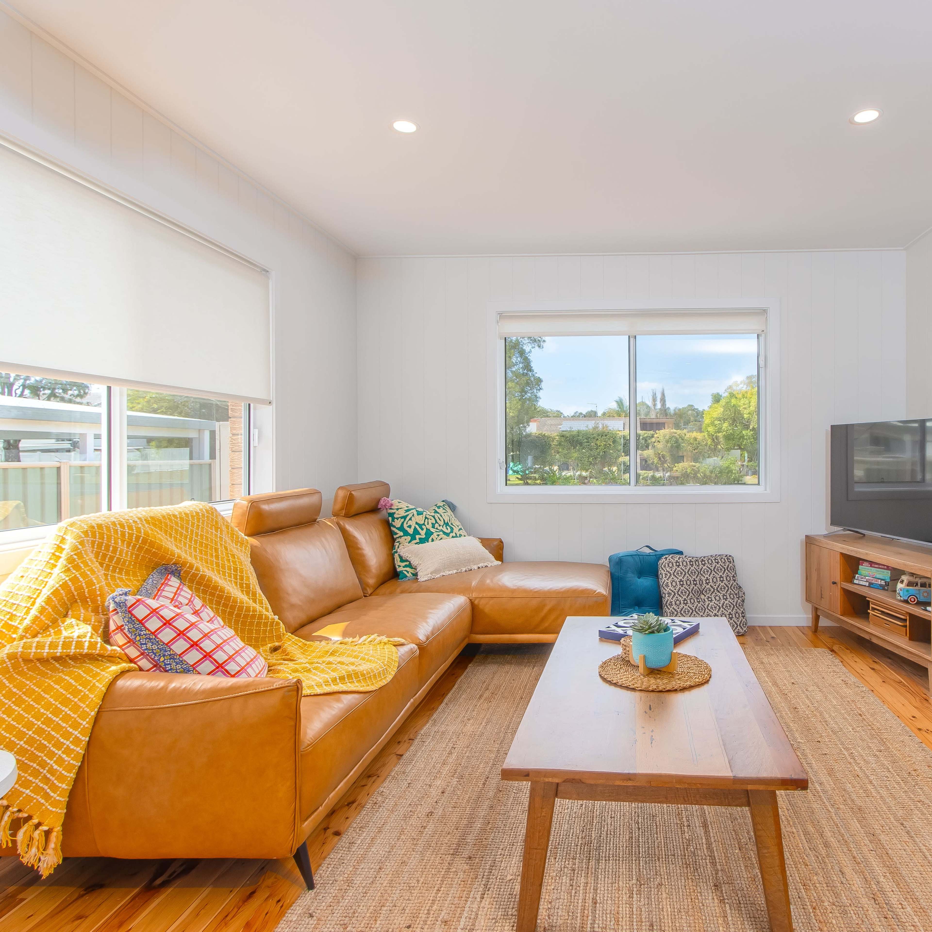 A cozy living room features a brown leather sofa, a wooden coffee table, and a television, with large windows providing a view of the outdoors.