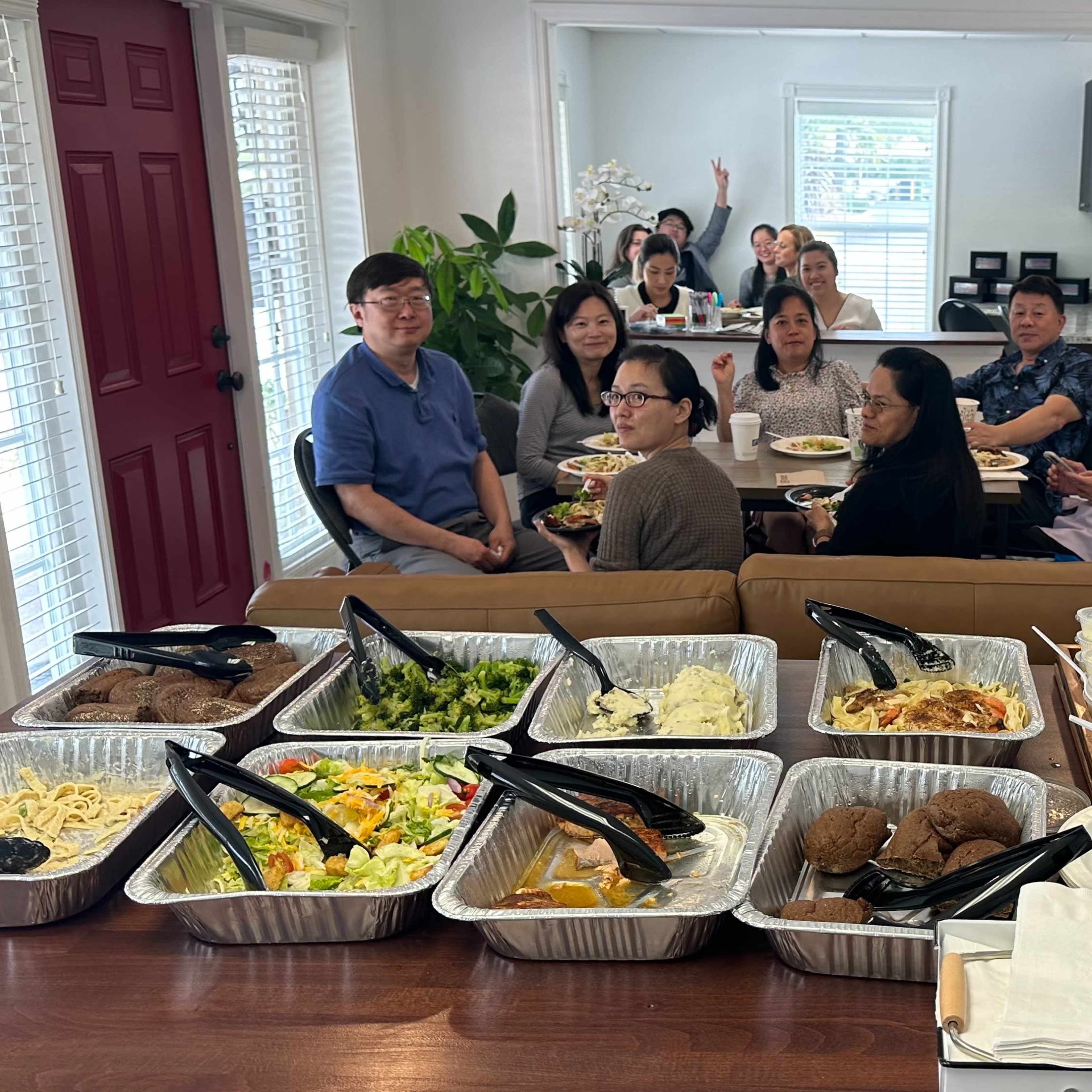 A group of people sits around a table enjoying a meal with various dishes displayed in aluminum trays in the foreground.