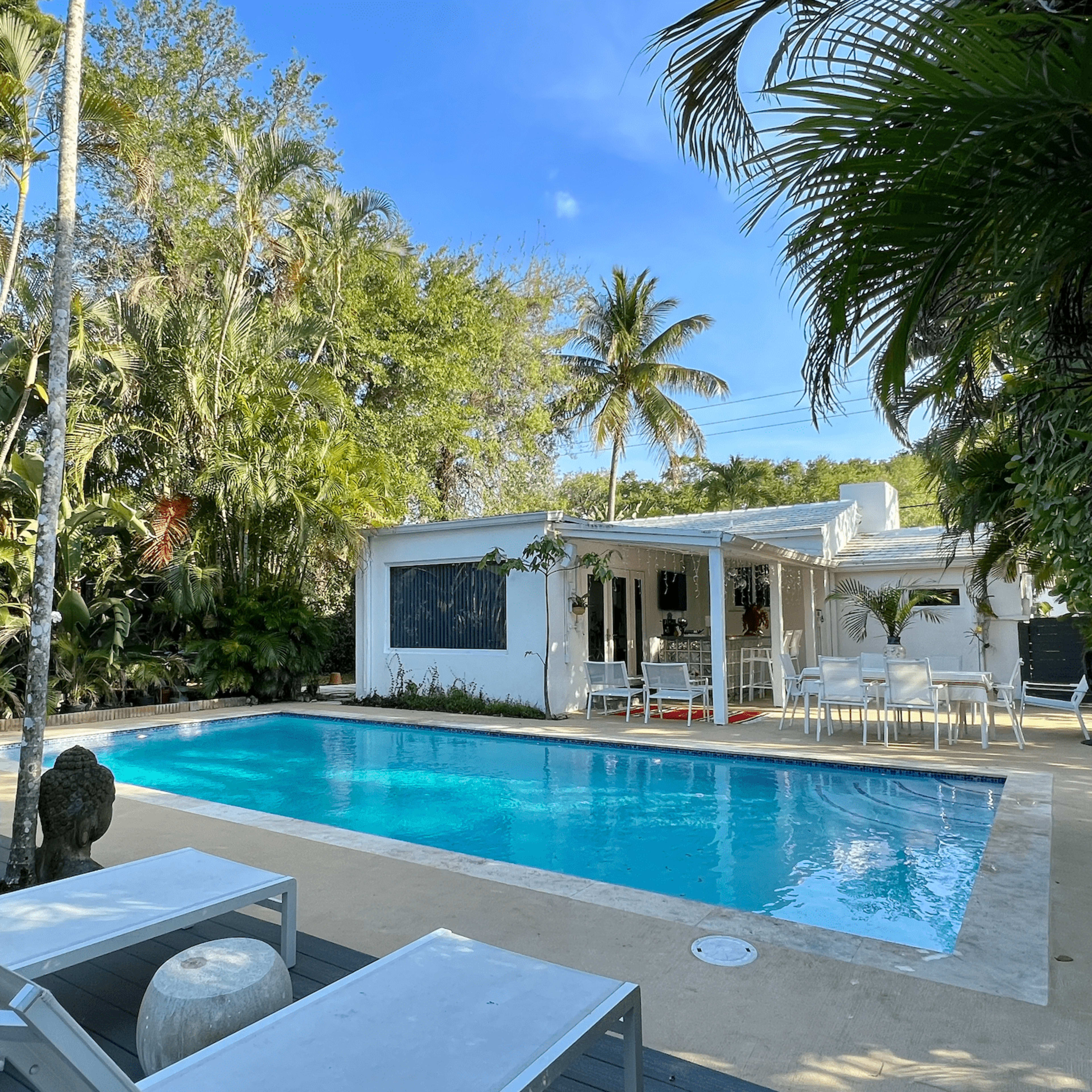 A swimming pool surrounded by lounge chairs and tropical foliage is located next to a white house with outdoor seating.