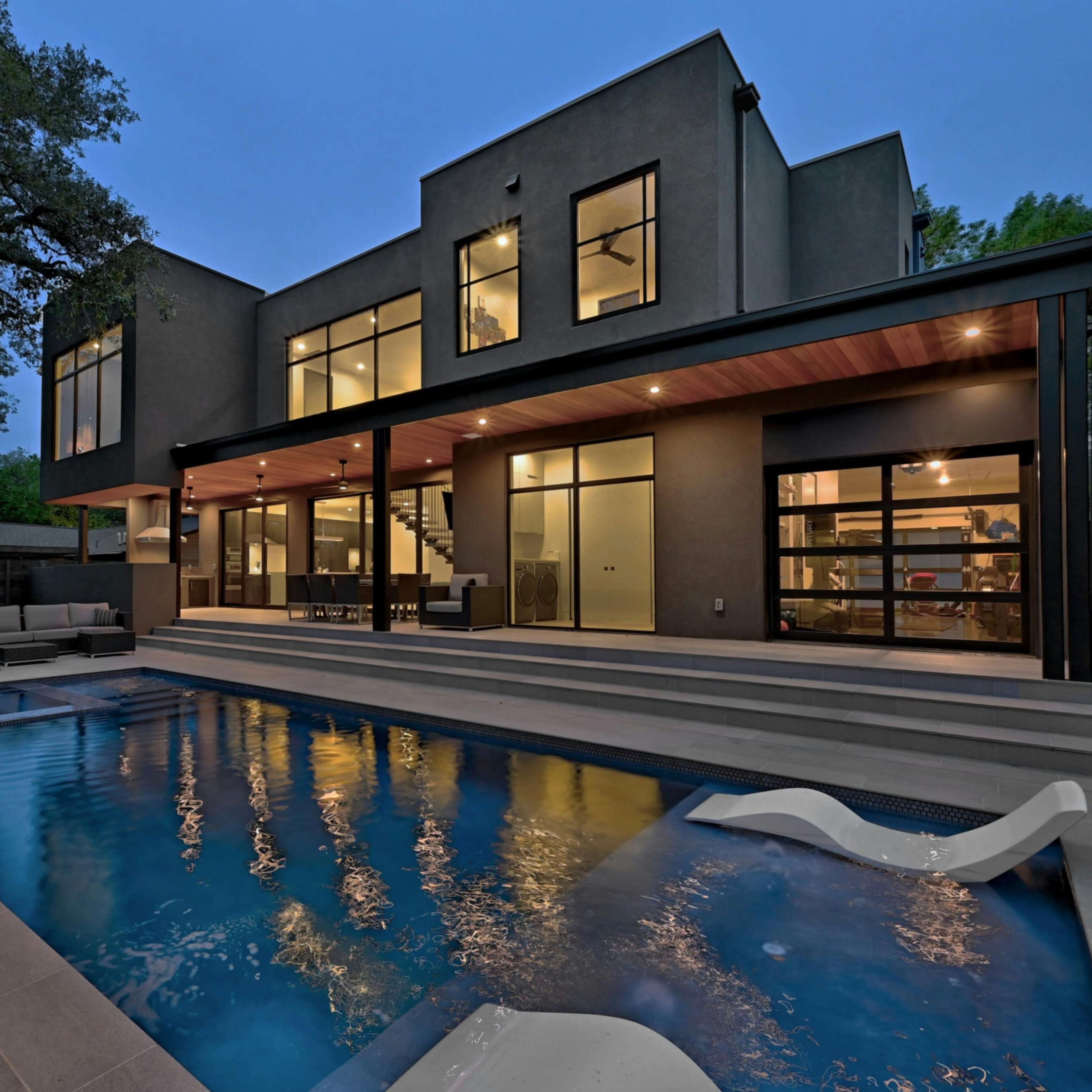 A modern two-story house with large windows overlooks a swimming pool surrounded by patio space, set against a twilight sky.
