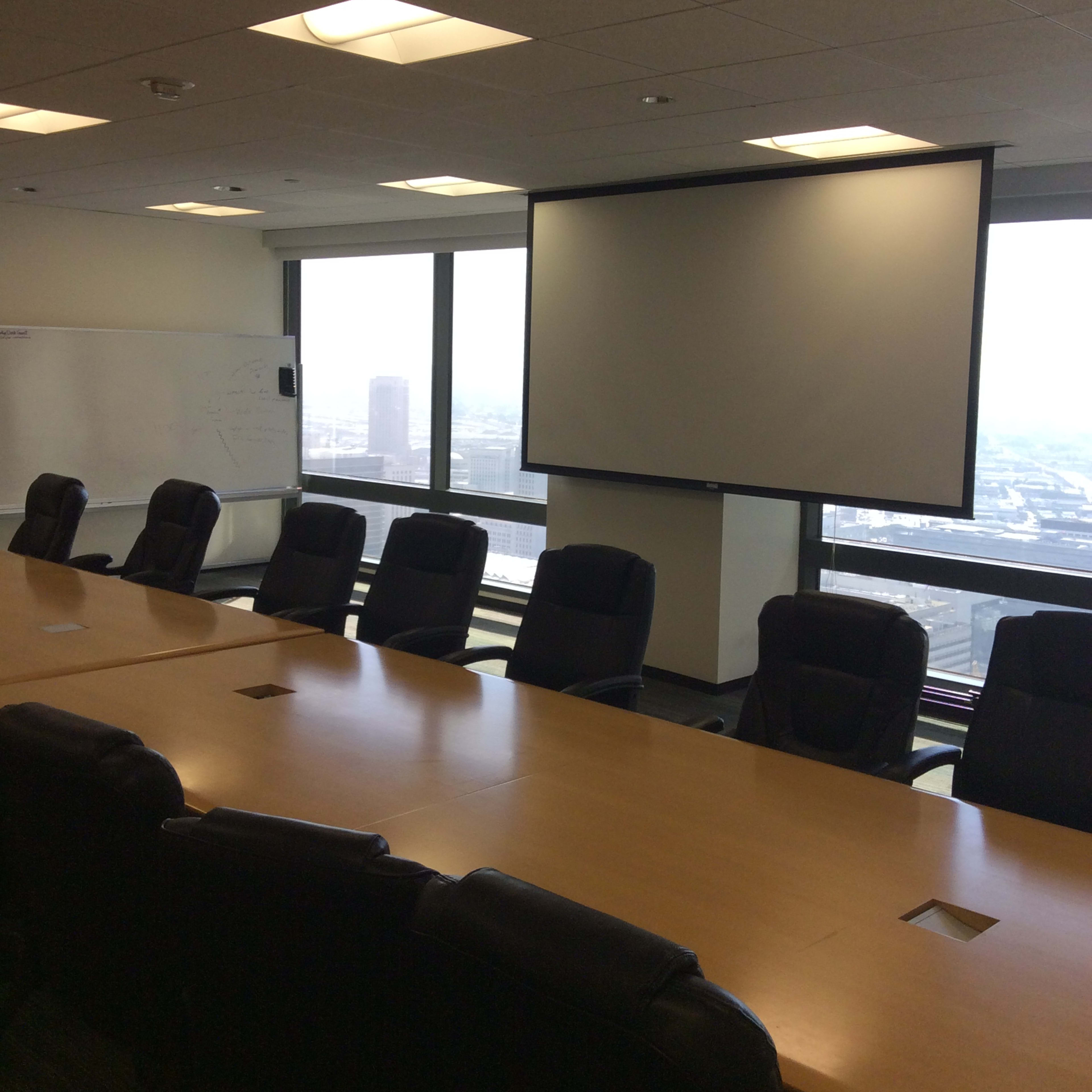 A conference room with a long wooden table and several black office chairs, featuring a large projector screen and windows overlooking a cityscape.